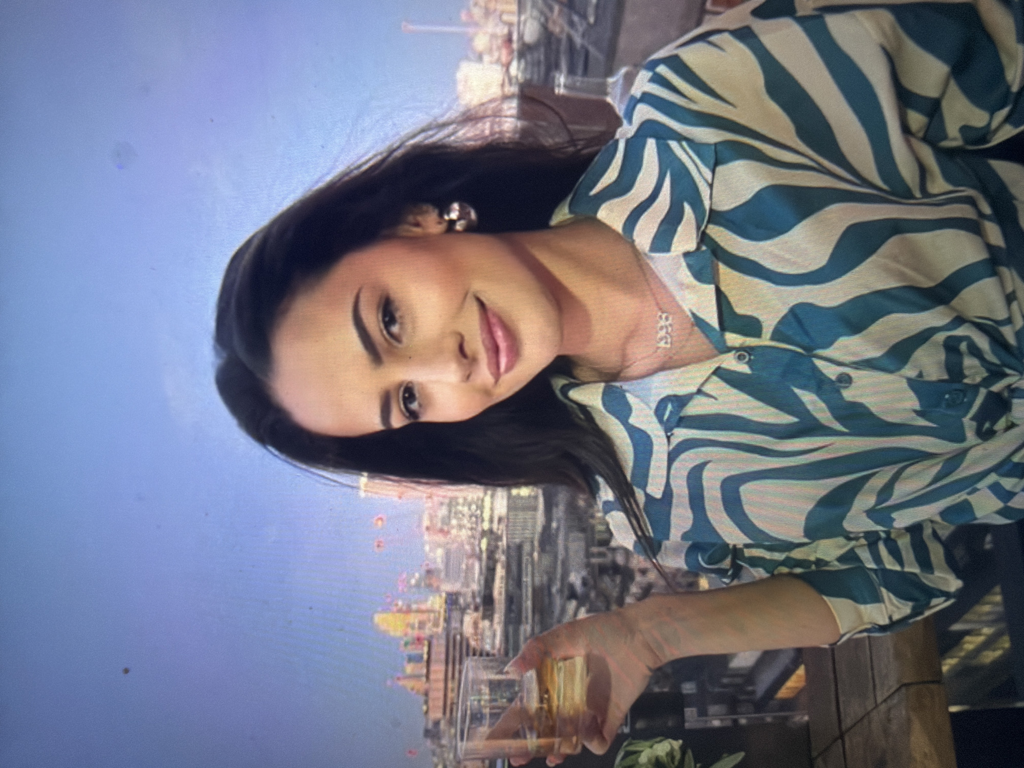 The image displays a young woman posing outdoors on a rooftop against a city skyline at dusk. The woman has dark, long hair, light-medium skin, and is looking directly at the camera with a subtle smile. She wears a long-sleeved, button-up shirt featuring a vibrant teal/blue and off-white/cream abstract stripe pattern, which appears tied at the front. Her left hand holds a clear glass tumbler containing a yellowish-amber liquid. She wears small, silver-toned hoop earrings and a silver-toned necklace with a pendant spelling "1998". The background features a blurred cityscape with numerous buildings and illuminated city lights, indicating evening. The sky above the city is a light blue, transitioning to a slightly darker hue. A dark table surface is partially visible to the woman's left. The specific location in the city of Shumen, Bulgaria, cannot be confirmed from the visual information presented in the image.