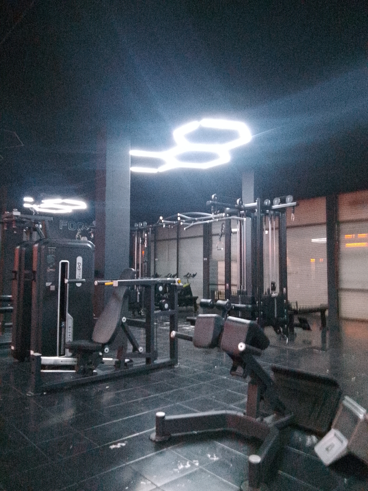 The image depicts an indoor gym with modern equipment and hexagonal LED lighting fixtures on the ceiling. The gym floor is dark and reflective, with visible lines. Several pieces of weight training equipment, including cable machines and a leg curl machine, are present. A large, dark grey machine with the partial word "FOO" visible on its side is in the foreground. The background shows more gym equipment and a large window with rolled-down blinds. The overall scene is dimly lit, with the bright LED lights creating reflections. The location is specified as Trenggalek, Indonesia. There are no people visible in the image.