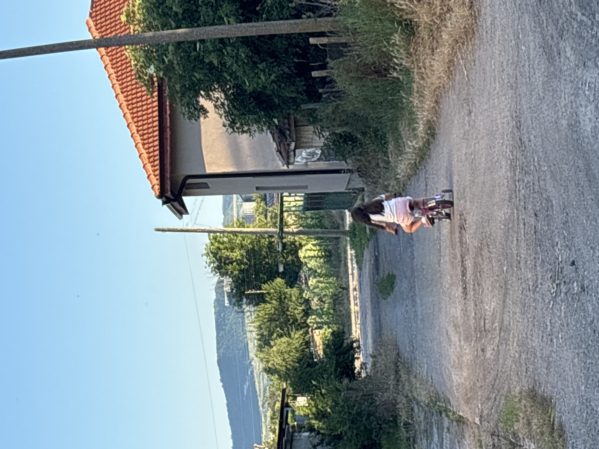 In Enevo, Bulgaria, a young girl with long dark hair, wearing a white t-shirt and pink shorts, rides a small pink bicycle with training wheels down a gravel road. The road is flanked by overgrown grass and bushes. To the right, a light-colored house with an orange tiled roof is visible, partially obscured by a large tree. A tall utility pole stands near the house. In the distance, rolling hills and more trees form the background under a clear blue sky. The scene appears to be a rural residential area during daylight.