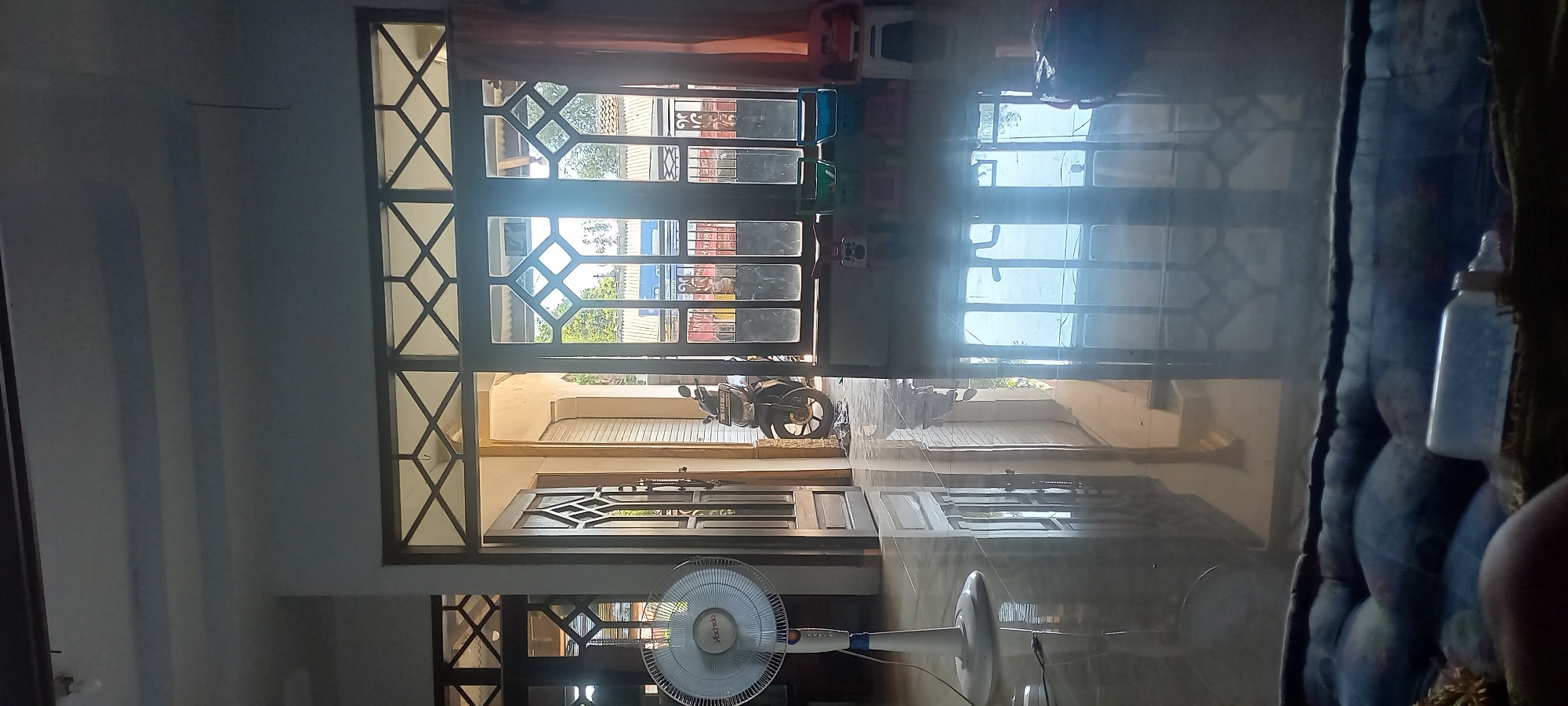 A wide-angle shot captures the interior of a house in Tongaswetan, Indonesia, facing outward through a large set of glass doors. The sunlit exterior, with glimpses of houses and trees, is visible beyond the doors. Inside, the polished floor reflects the light and the geometric patterns of the door frames. A white electric fan stands to the left, its base on the floor. To the right of the fan, a wooden door is ajar, revealing a motorcycle parked in what appears to be an entryway or adjacent room. Further inside, behind the motorcycle, a rolling metal door is visible. The scene is calm and appears to be captured during daylight hours. The foreground is occupied by a partially visible cushioned surface, possibly a mattress or sofa, with a plastic bottle on it.