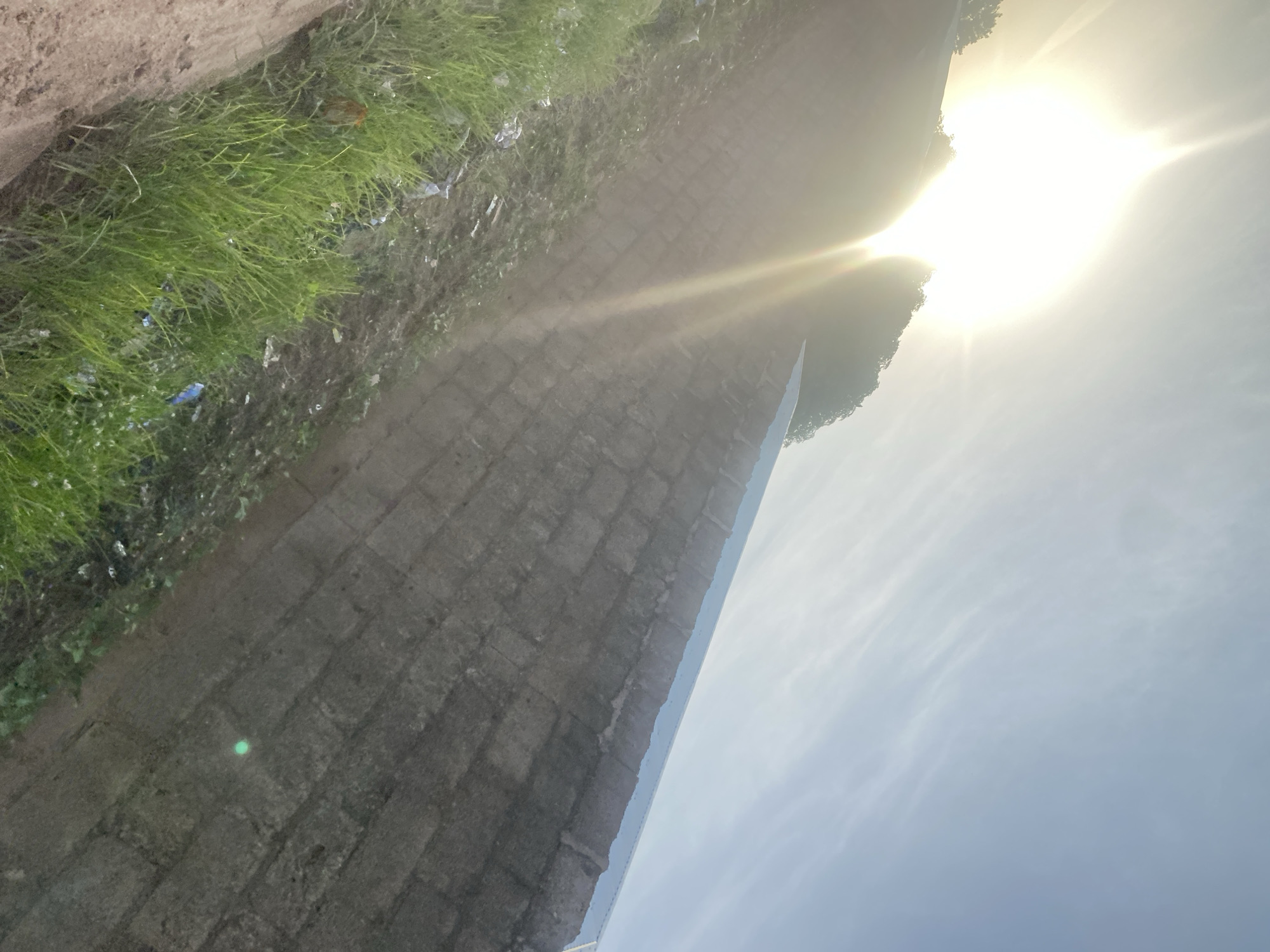 The image depicts a bright, sunny sky with a low-lying sun creating a prominent lens flare. In the foreground, there is a weathered brick wall and a patch of green grass with some debris scattered within it. The perspective suggests the photo was taken at ground level, looking upwards towards the sky and adjacent embankment. The brick wall appears to be part of a pathway or structure. The overall scene suggests an outdoor environment, possibly an urban or semi-urban area. The specific location in Jalingo, Nigeria, is not discernible from the visual elements of the image itself.