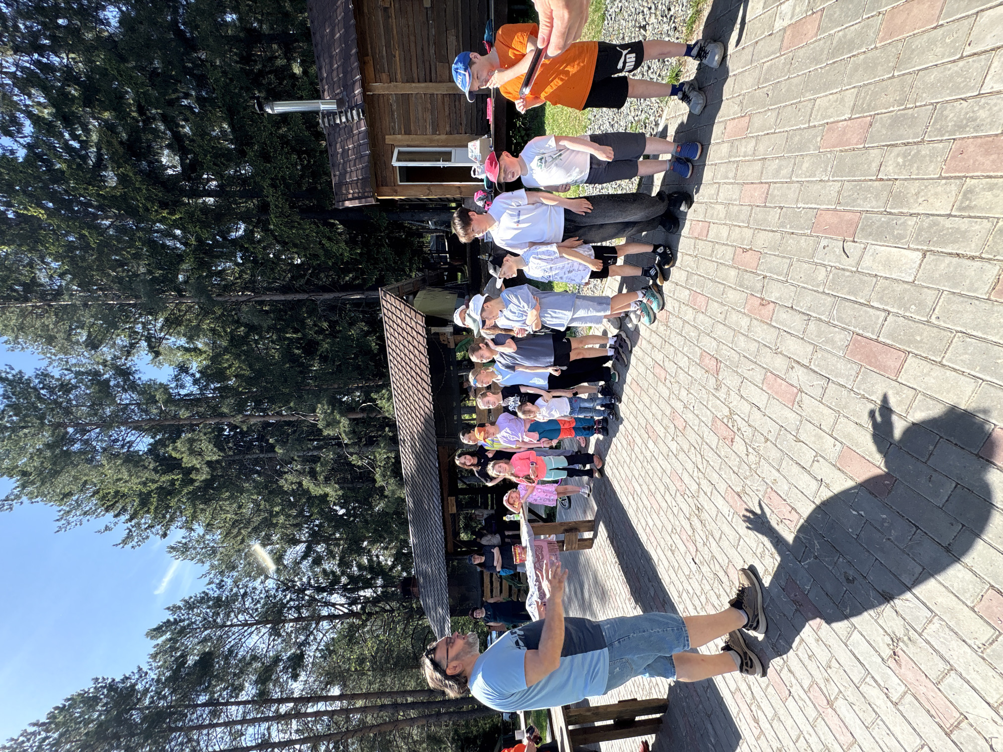 The image shows a group of children standing in a line on a brick-paved area, likely outdoors. An adult male is standing in front of them, possibly instructing or addressing them. The location appears to be near a wooden building and surrounded by trees, based on the architecture and vegetation, the location is near the city of Прикамский, Russia. The group of children consists of a variety of ages and are wearing diverse casual clothing. The adult male is wearing a light blue t-shirt, sunglasses, and denim shorts. The lighting suggests it is daytime, with the sun casting shadows on the ground.