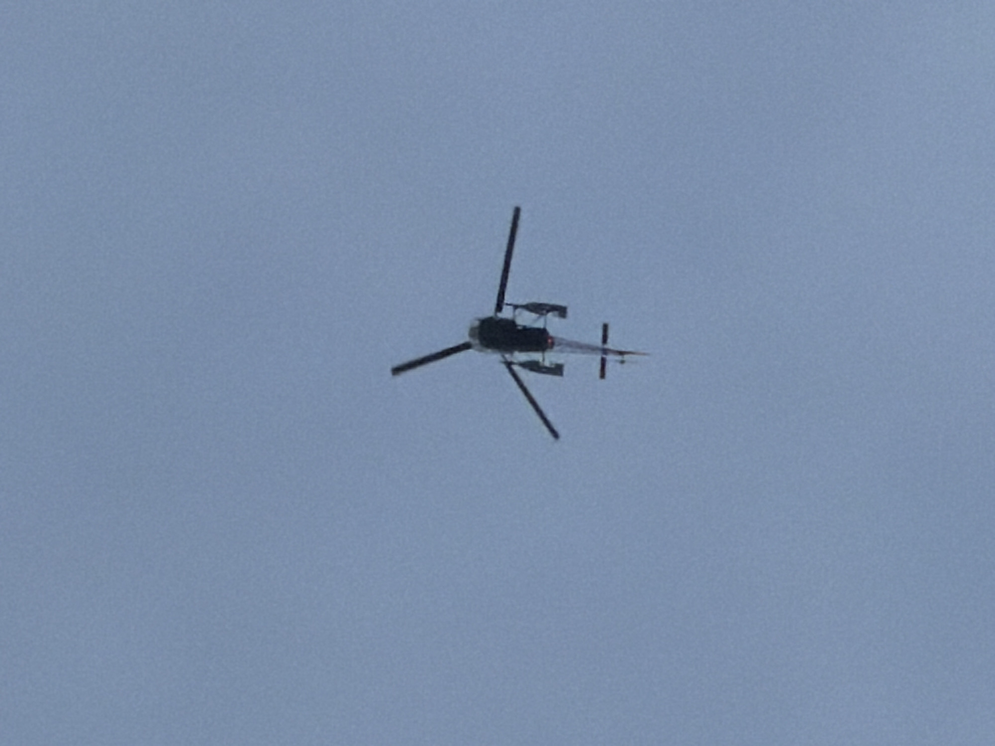 A black helicopter is captured in flight against a pale blue, overcast sky. The helicopter is viewed from below, with its main rotor blades blurred due to motion. The tail rotor is visible, as are the landing skids. The helicopter's body is dark, with a small red or orange accent visible on the tail boom. The overall impression is of a clear, unobstructed view of the sky. The image does not contain any identifiable elements of the city of Leh, India.