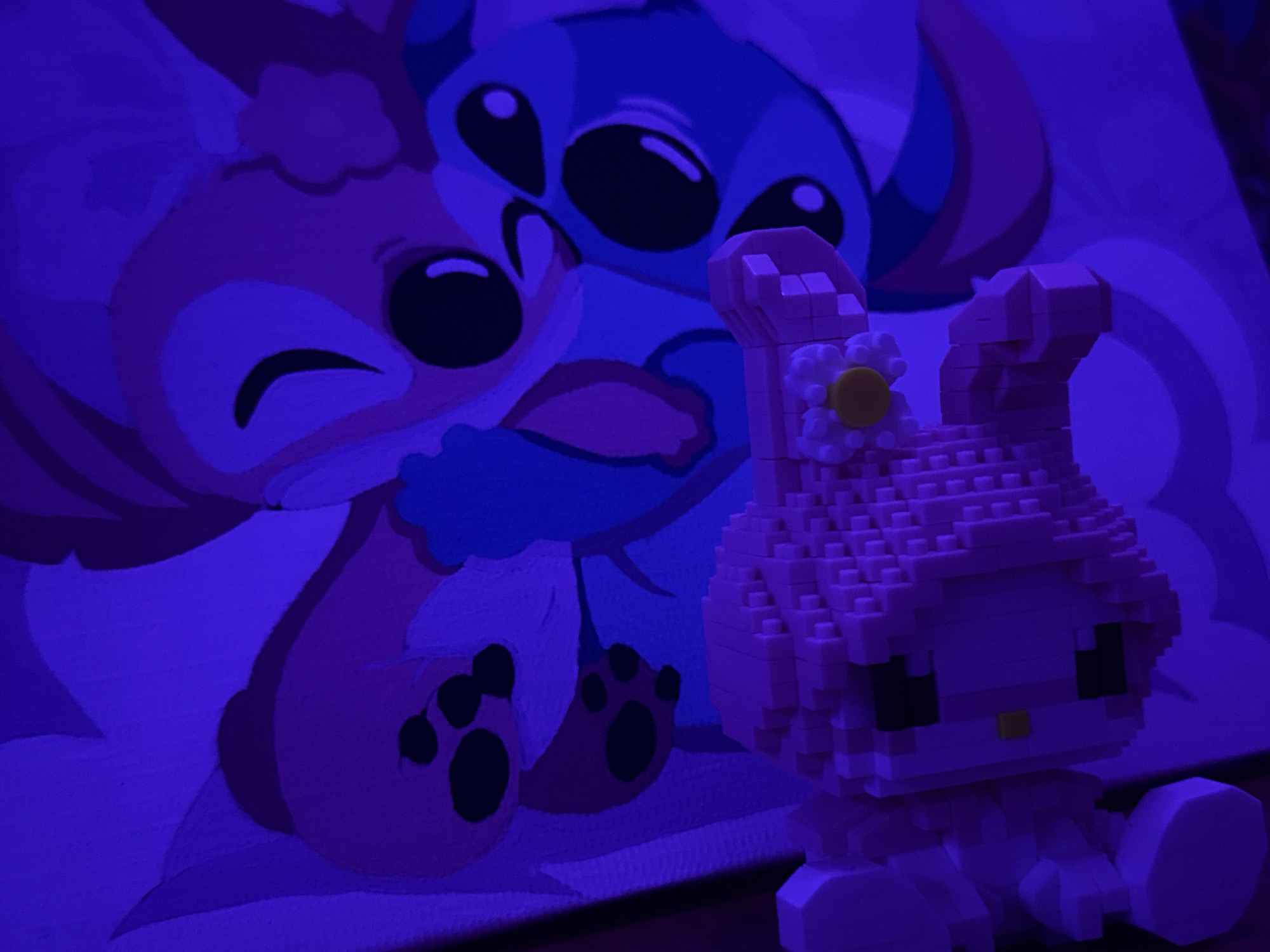 In a dimly lit room illuminated by purple light, a painting of two cartoon characters, resembling Stitch and Angel from Lilo & Stitch, is displayed. The characters are depicted embracing each other. In the foreground, a pink, block-style figurine of Hello Kitty sits with a yellow flower on its head. The figurine is positioned to the right of the painting. The scene appears to be indoors, likely in a residential setting in Rayevskiy, Russia, although this specific location cannot be confirmed from the image alone.