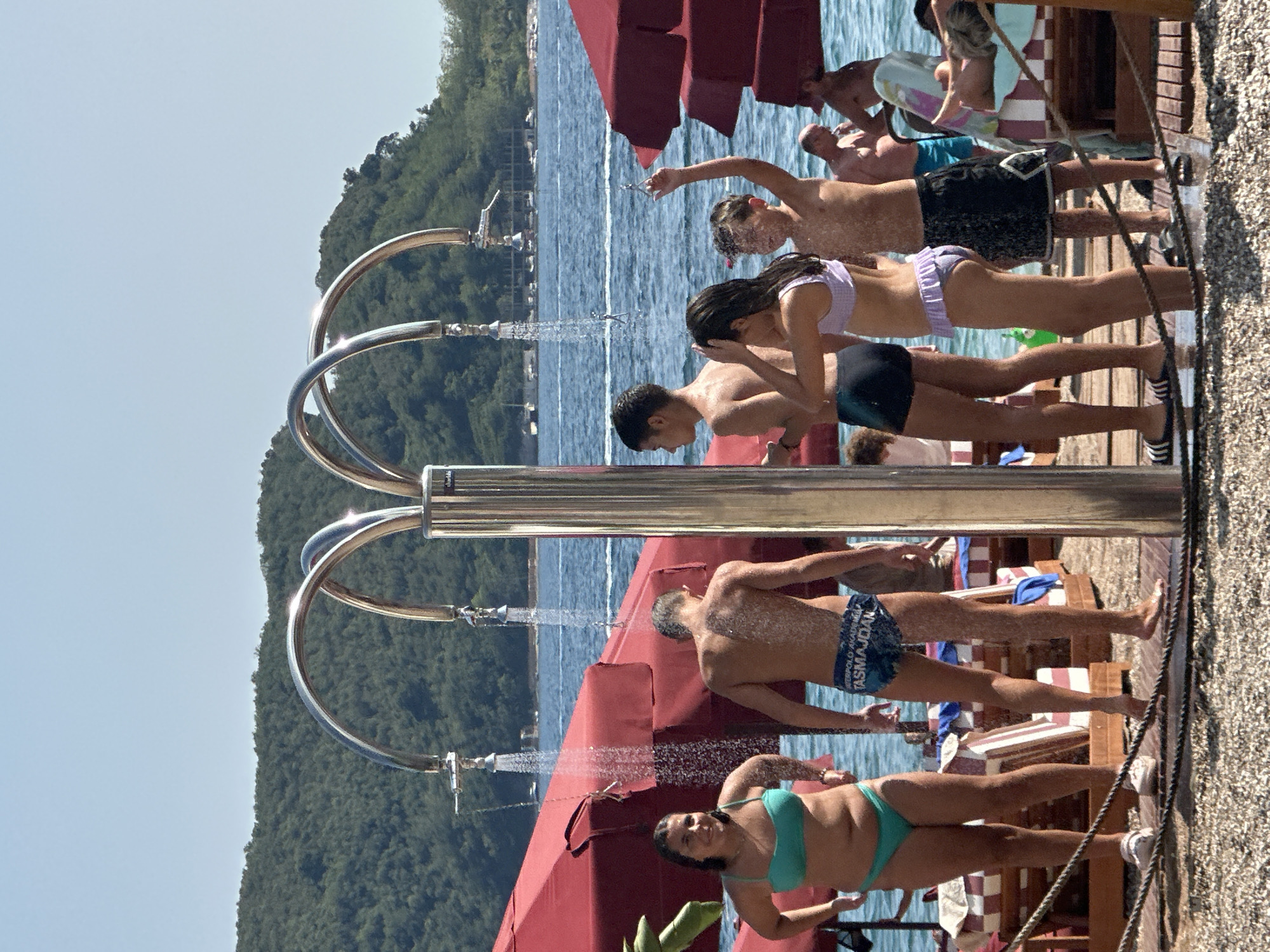 A group of people are showering outdoors on a sunny day, near a body of water. The shower station appears to be on a beach or promenade, with a paved area in the foreground and lounge chairs and umbrellas set up in the background. The water is running from the shower heads, and several people are standing under them, getting wet. The background shows a calm blue sea, with a tree-covered hill in the distance, suggesting a coastal location. This scene is consistent with the beaches of Budva, Montenegro, known for their clear waters and scenic beauty.