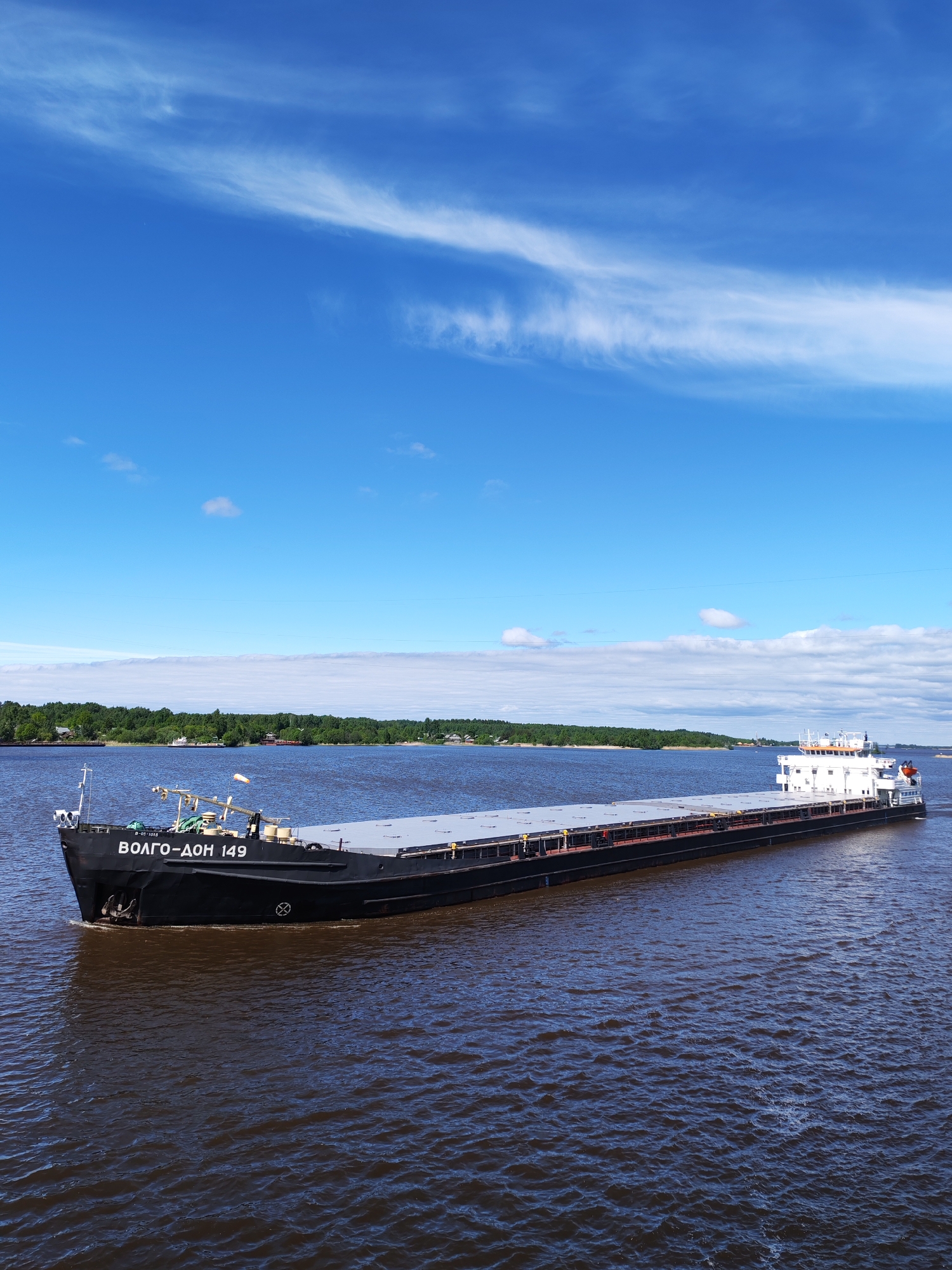 A long, black cargo barge labeled "ВОЛГО-ДОН 149" is traveling across a large body of water. The sky above is bright blue with streaks of white clouds. The water is dark and choppy. A green, forested shoreline is visible in the background. The barge is moving from left to right. The location is Воднема, Russia.