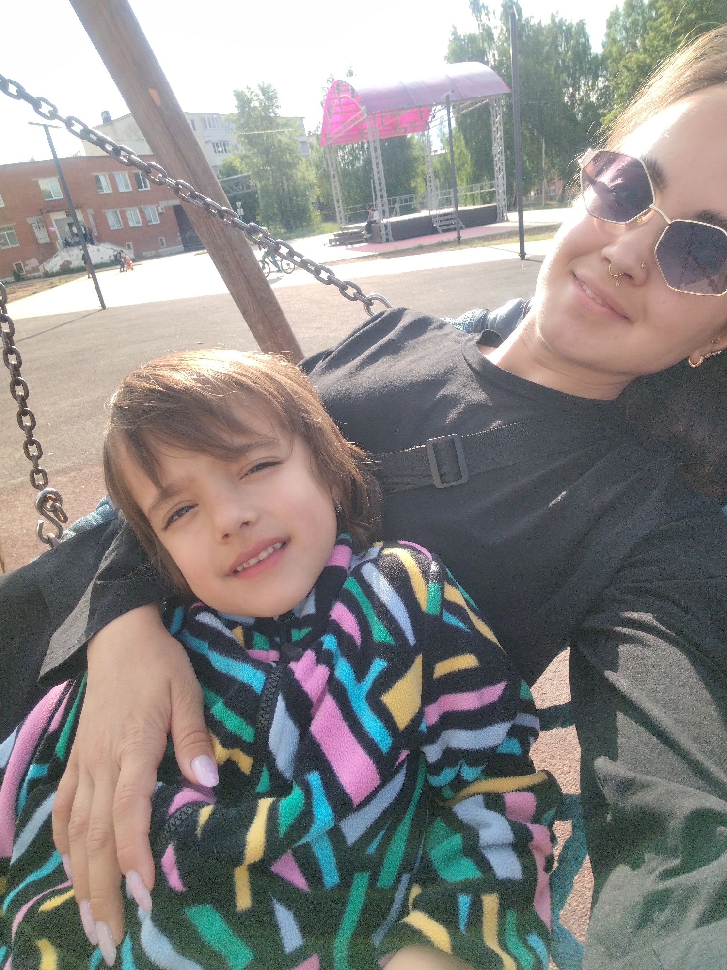 The image shows a woman and a child on a swing set in what appears to be a park in Noviy Urengoy, Russia. The woman is wearing sunglasses, a black long-sleeved shirt, and has a nose ring. The child is wearing a colorful fleece jacket with a geometric pattern. The swing set is made of wood and metal chains. In the background, there is a pink gazebo-like structure, a brick building, and trees. The ground is paved. The woman is holding the child, and both are facing the camera.