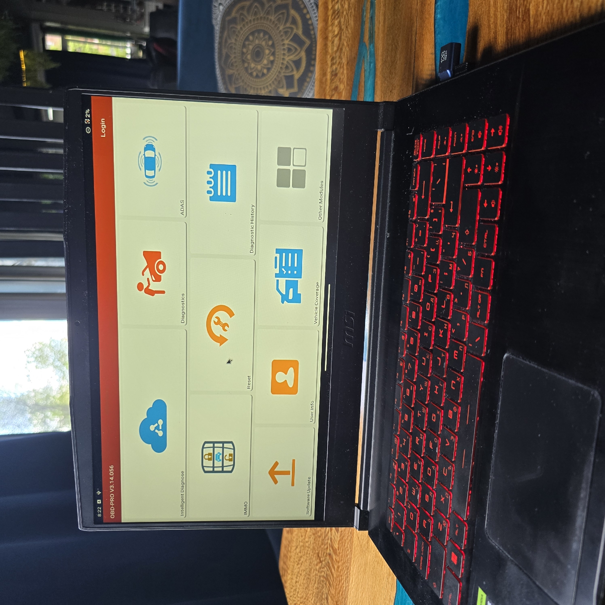 The image shows a black laptop, likely an MSI model due to a visible logo on its hinge, open and displaying a specialized software interface. The laptop has a red backlit keyboard, and a small black USB 3.1 flash drive, labeled "128 GB," is plugged into its right side.
The screen displays the graphical user interface for an automotive diagnostic application identified as "OBD-PRO V3.14.056." The current time on the screen is "8:22," and the battery charge is critically low at "2%." The software features a grid of icons and labels for various functions, including "Login," "Intelligent Diagnose" (represented by a cloud with gears), "IMMO" (a grid with a lock and car), "Software Update" (an upward arrow), "User Info" (a person icon), "Reset" (a circular arrow with a wrench), "Diagnostics" (a person with a car engine), "ADAS" (a car with radar waves), "Diagnostic History" (a document icon), "Vehicle Coverage" (a document with a car symbol), and "Other Modules" (a grid of squares).
The laptop rests on a wooden surface. In the background, through a window, out-of-focus trees and building structures are visible, indicating an indoor setting during daylight hours, possibly morning or evening. A blue decorative mat with a circular pattern is also partly visible in the background. The scene depicts a setup for vehicle diagnostics, taken in Sofia, Bulgaria.