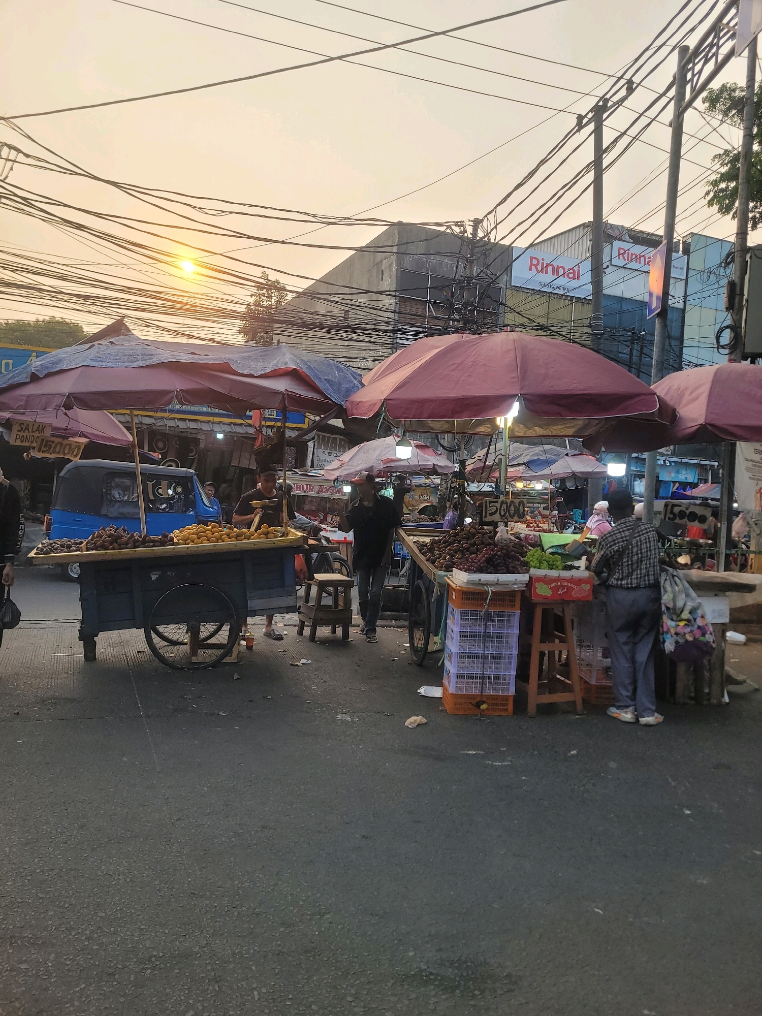 This image captures a street scene in South Jakarta, Indonesia, likely during the late afternoon or early evening, as indicated by the warm glow of the setting sun and the presence of streetlights. The foreground features a bustling roadside market with several vendors selling fruits and other goods from carts and stalls.
Dominating the left side is a fruit cart laden with produce, possibly sweet potatoes and corn. Behind it, a blue vehicle, possibly a small truck or van, is parked. Two individuals are interacting near this cart. Further to the right, another vendor operates from a stall covered by a large red umbrella, showcasing a variety of fruits, including grapes, and other items. A person in a checkered shirt is attending to this stall, and another person in jeans stands nearby. The scene is characterized by a dense network of overhead electrical wires, a common sight in urban Indonesian settings. In the background, commercial buildings are visible, one with a prominent "Rinnai" sign. The ground is paved with asphalt, and there are scattered pieces of trash and debris. The overall atmosphere suggests a vibrant and active marketplace.