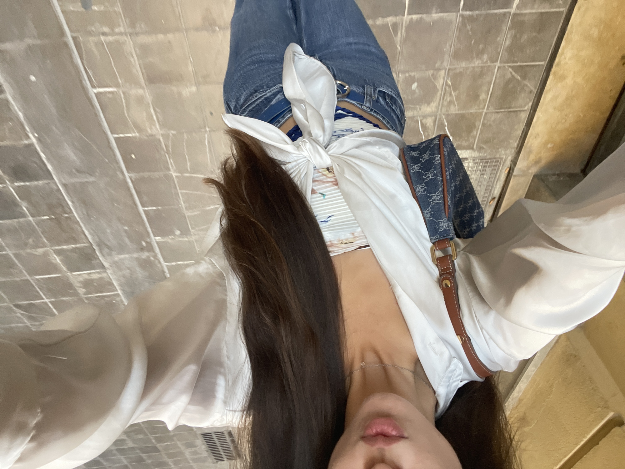 Here is a description of the image:
The image depicts a person standing on a tiled surface, possibly a sidewalk. The person is wearing jeans, a white tied shirt and a denim handbag with brown straps. The location is possibly València, Spain. The person is facing the camera, capturing an upward angled shot of their torso and legs. The background is a tiled surface and an adjacent wall.