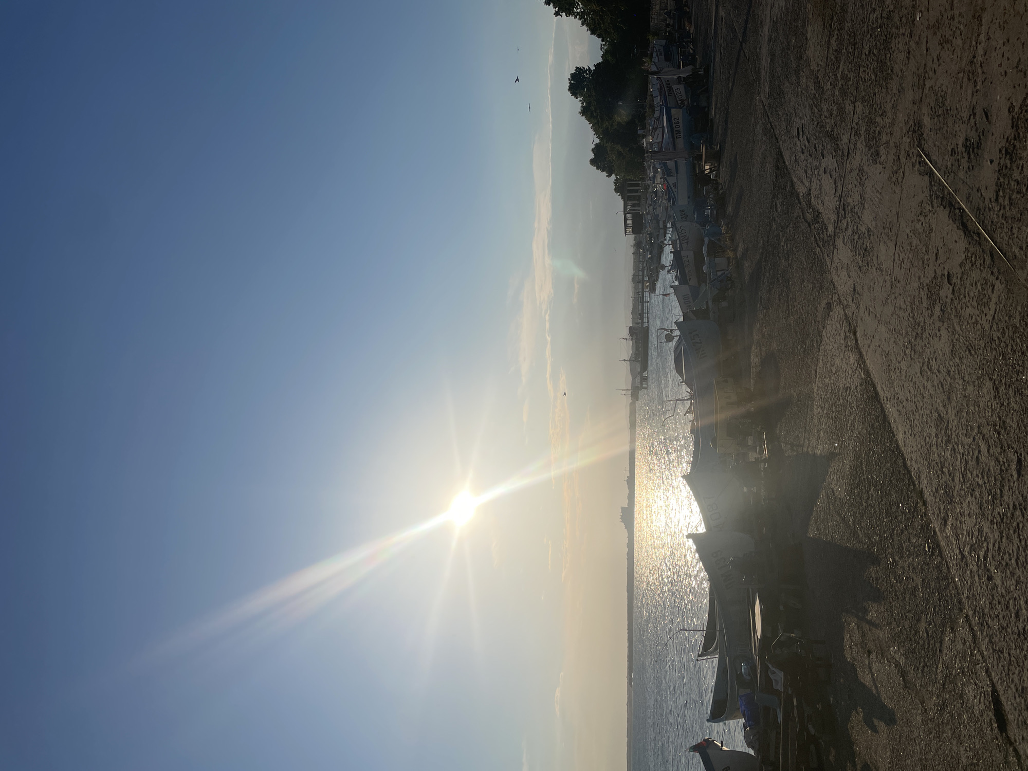 A high-angle, eye-level shot captures the Bulgarian city of Pomorie, specifically its coastline and harbor area, during what appears to be sunset. The sun, a bright orb with prominent light flares, dominates the upper left quadrant of the frame, casting a shimmering golden path across the calm, rippling sea. The water stretches towards a distant, hazy horizon, where a faint silhouette of industrial structures and landmass is visible.
Along the shore, a collection of small fishing boats rests on the sandy, gravelly ground. Many are lined up parallel to the water's edge, with some partially submerged. The boats are diverse in design and condition, some appearing well-maintained, others weathered. Several boats have visible identification numbers or markings, such as "№001," "№062," and "№023." Structures that appear to be part of the harbor infrastructure, including wooden docks and small buildings with covered roofs, are scattered amongst the boats.
The shoreline itself is a mix of dark, coarse sand and scattered debris. Dense green trees line the far right of the frame, forming a backdrop to the harbor scene. The sky above is a clear, gradient blue, transitioning to lighter hues near the horizon. A few wispy clouds are present, catching the golden light of the setting sun. Two birds are visible in flight, adding a sense of life to the serene landscape. The overall impression is one of a peaceful, working harbor at the close of day.