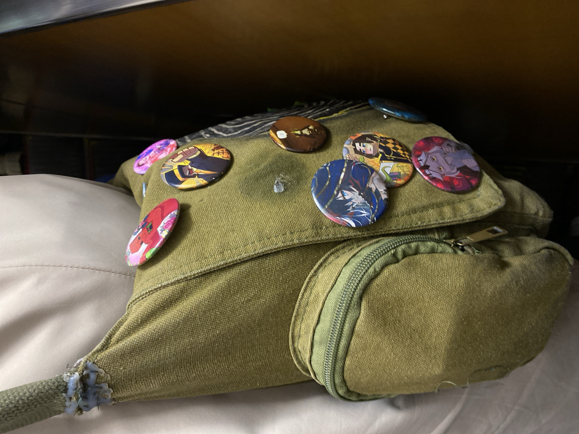 A green canvas bag sits on a bed or other soft surface, likely indoors under a shelf. The bag is decorated with multiple round, colorful pins depicting anime-style characters. The bag has a flap that covers a zipped pocket. The location is unknown and there is no evidence to suggest this image was taken in Saint Petersburg, Russia.