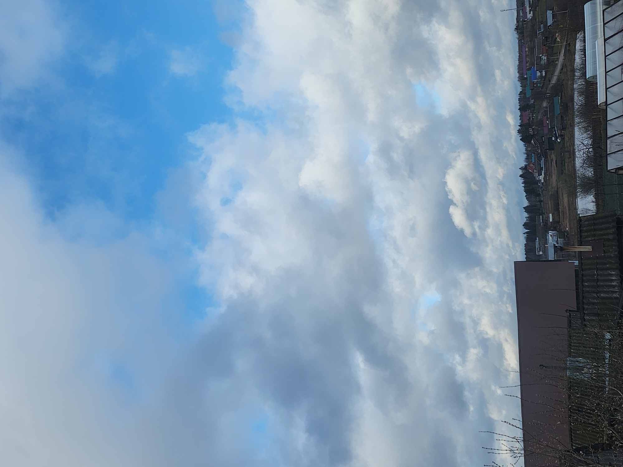 The image depicts a partly cloudy sky over a residential area, likely in Krasnogorskoye, Russia. The sky is a mix of blue patches and thick, grey clouds. In the distance, houses are visible along the horizon. Closer to the viewer, there is a building or structure with a brown exterior. Bare tree branches are visible in the foreground. A greenhouse is visible next to the houses along the horizon.