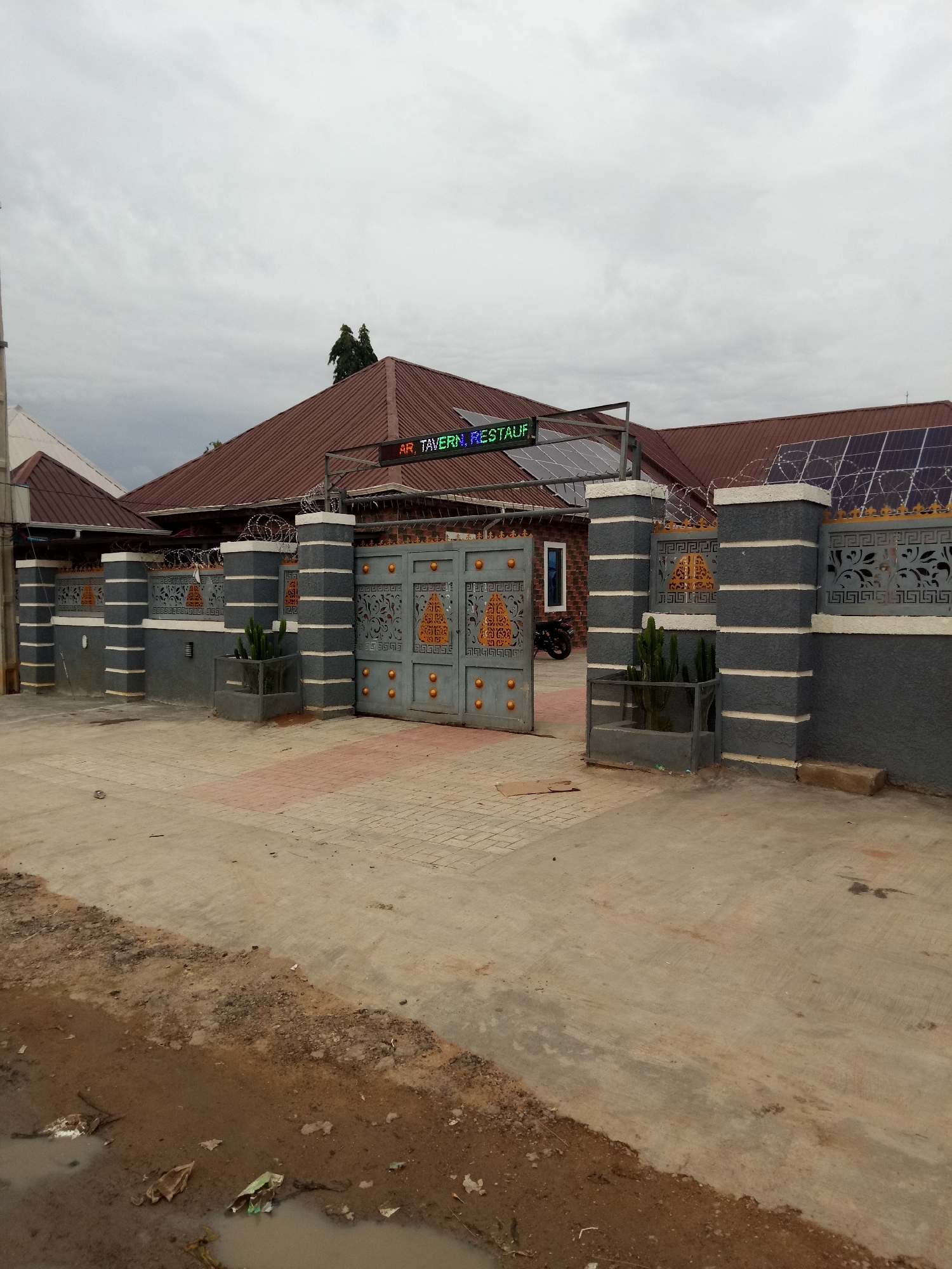 The image depicts the entrance of a establishment identified as "AR. TAVERN. RESTAUF." located in Uke, Nigeria. The entrance features a large, ornate metal gate with golden circular accents and a repeating geometric pattern. The gate is flanked by grey pillars with white bands, topped with decorative railings and barbed wire. Beyond the gate, solar panels are visible on the brown, sloped roof of a brick building. The surrounding area is paved with interlocking bricks and concrete, with some patches of mud and scattered debris in the foreground. Two potted plants, resembling cacti, are placed near the gate. A motorcycle is partially visible inside the compound to the left of the entrance. The sky is overcast.