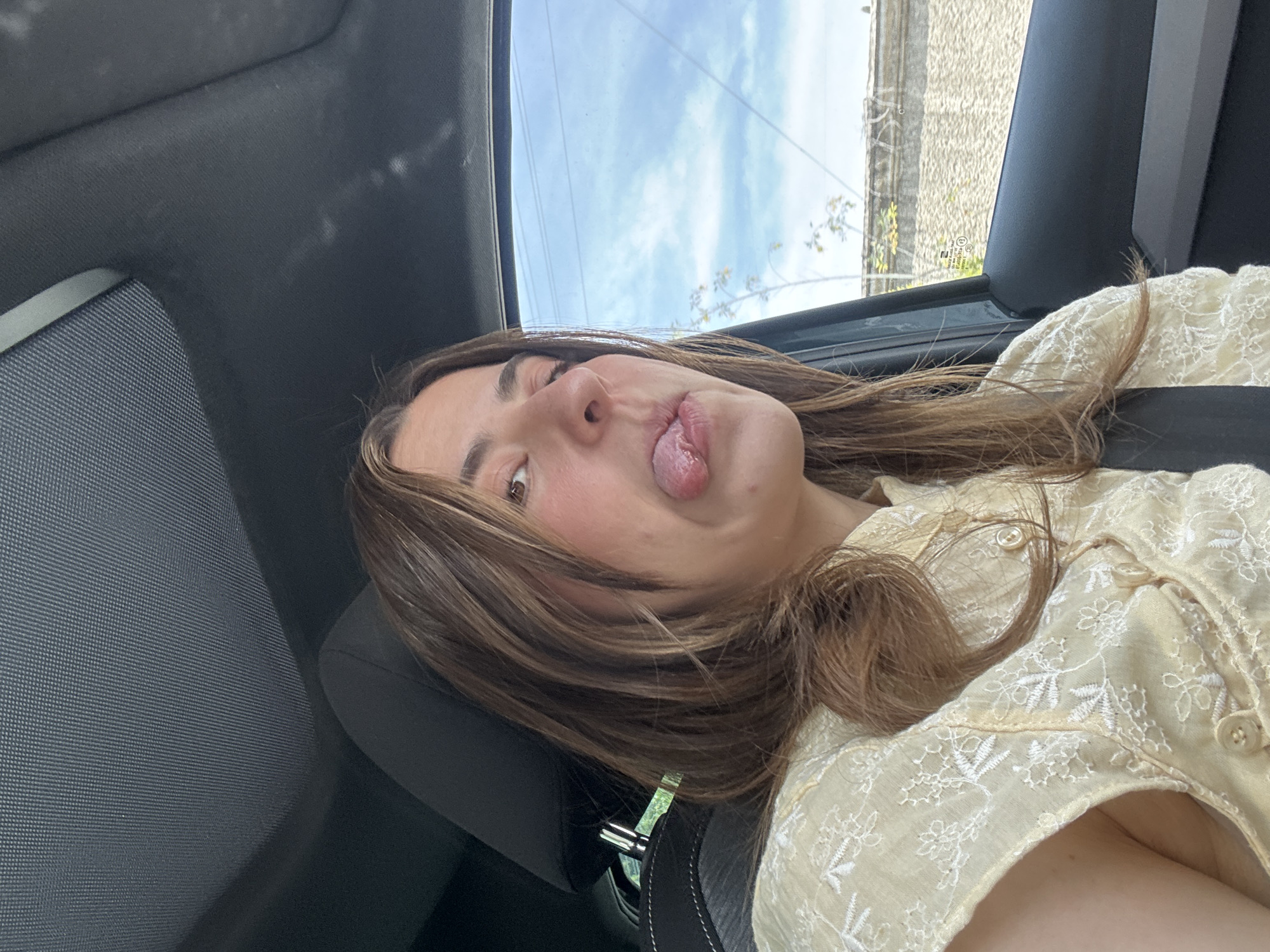 A woman is inside a car, sticking her tongue out. She has medium-length brown hair and is wearing a light yellow embroidered shirt. She is wearing a seatbelt. The background visible through the car window shows a blue sky, some greenery, and a stone wall. The location cannot be determined specifically to Puebla, Mexico based solely on this image.