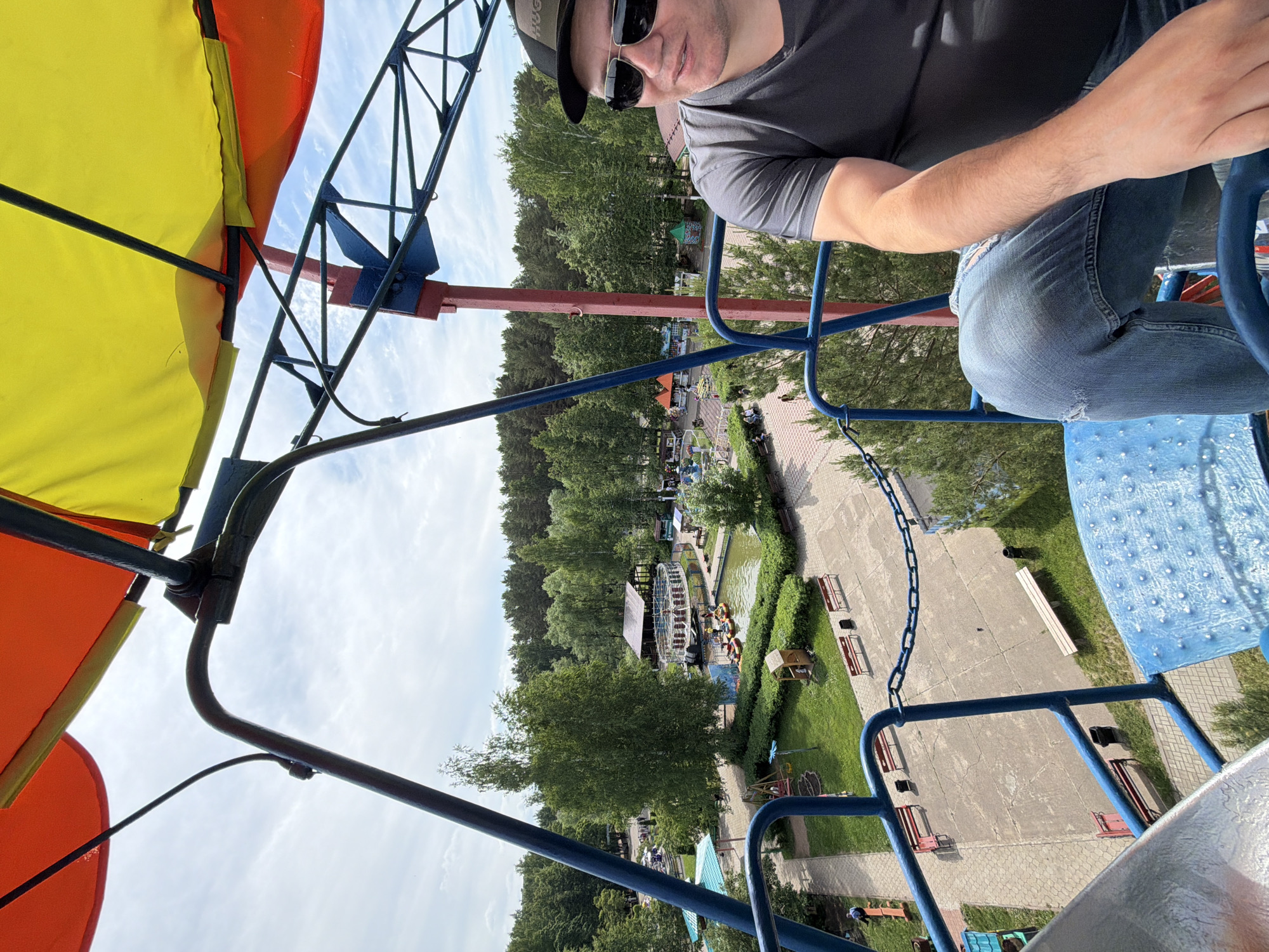 The image, taken from a high vantage point, likely an amusement park ride, captures a man and the park scenery below. The primary subject is a man in the foreground, seen from the chest up, wearing a dark grey t-shirt, blue jeans, sunglasses, and a black baseball cap with white text that appears to read "HUG" on the front. His left arm is visible, resting on a blue metal bar of the ride.
The ride itself features prominent yellow and orange fabric canopies, supported by a black metal framework. Blue metal railings and a chain are also part of the ride's structure. The man is seated in what appears to be a carriage or gondola, with a patterned blue surface visible to his right, likely part of the seating.
Below, an amusement park or recreational area in Chaikovsky, Russia, unfolds. Lush green trees dominate the landscape, interspersed with paved paths, grass areas, and various park attractions. A central feature is a body of water, possibly a pond or canal, where several people are seen in small boats, some with orange buoyancy aids, near a water wheel or similar attraction. Benches are scattered along the paths, and several small buildings and structures are visible throughout the park. The sky is bright with scattered white clouds, indicating a clear, sunny day, likely during the late morning or afternoon. The overall scene suggests a pleasant day out at an active amusement park.