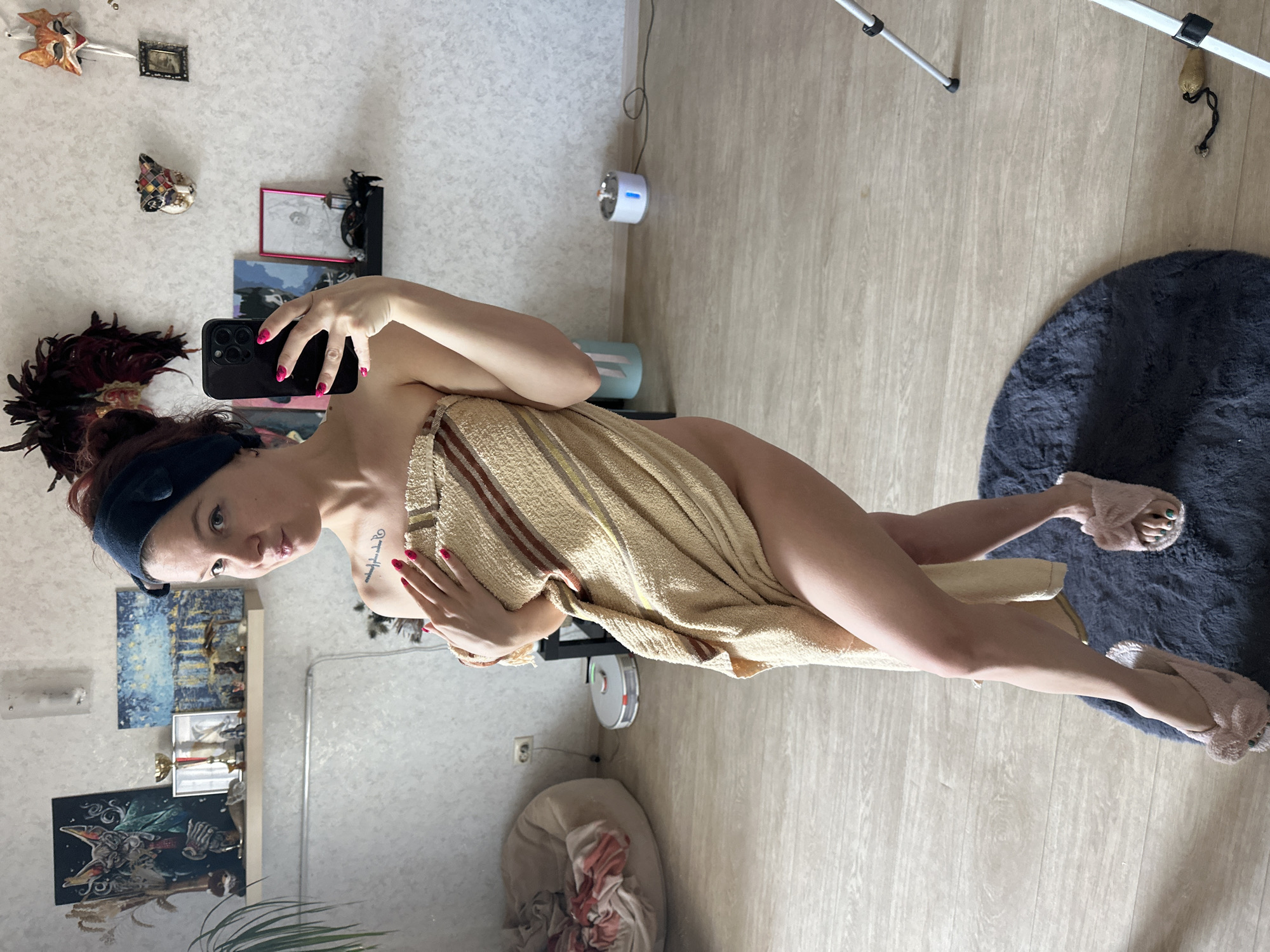 The image depicts a young adult female taking a mirror selfie within a residential interior in Vladimir, Russia. The woman, positioned centrally, is wrapped in a beige and brown striped towel, covering her torso and extending down her upper thighs. She wears light pink fluffy slippers, revealing green nail polish on her toenails. Her left hand holds a black smartphone with multiple camera lenses, aimed towards herself. Her right hand rests over the towel at her chest. She has dark hair styled back with a dark blue head wrap, and a small script tattoo is visible on her right shoulder/collarbone area. Her lips are slightly pursed, and she wears light eye makeup.
The flooring consists of light wood-look planks, with a circular dark grey shag rug placed directly beneath her feet. The walls are light-colored with a subtle textured finish.
To the left of the subject, a large, light beige beanbag chair is visible, piled with clothing or fabric in shades of pink and white. Above the beanbag, a white electrical outlet is mounted on the wall. On the floor nearby, a white robotic vacuum cleaner is present. Above these elements, a white floating shelf displays various decorative items: a vase containing dried plant material, a dark painting depicting a stylized animal (possibly a cat or fox) in a green suit, a small golden trophy, a framed picture, another golden trophy, and a blue-toned impressionistic painting. A large green houseplant is partially visible on the far left edge of the frame.
To the right of the subject, the wall is decorated with several items: a large feathery dark-colored headpiece or mask, a smaller harlequin-patterned mask, and a white and orange fox-like mask from which a white ribbon hangs. Below these, a black floating shelf holds a small picture in a pink frame and a miniature decorative lantern. On the floor to the right, a silver and white pet water fountain with a blue indicator light is visible, connected by a black power cord. Further to the right, a silver-colored tripod with black accents is partially visible, with a small golden pouch hanging from one of its legs.