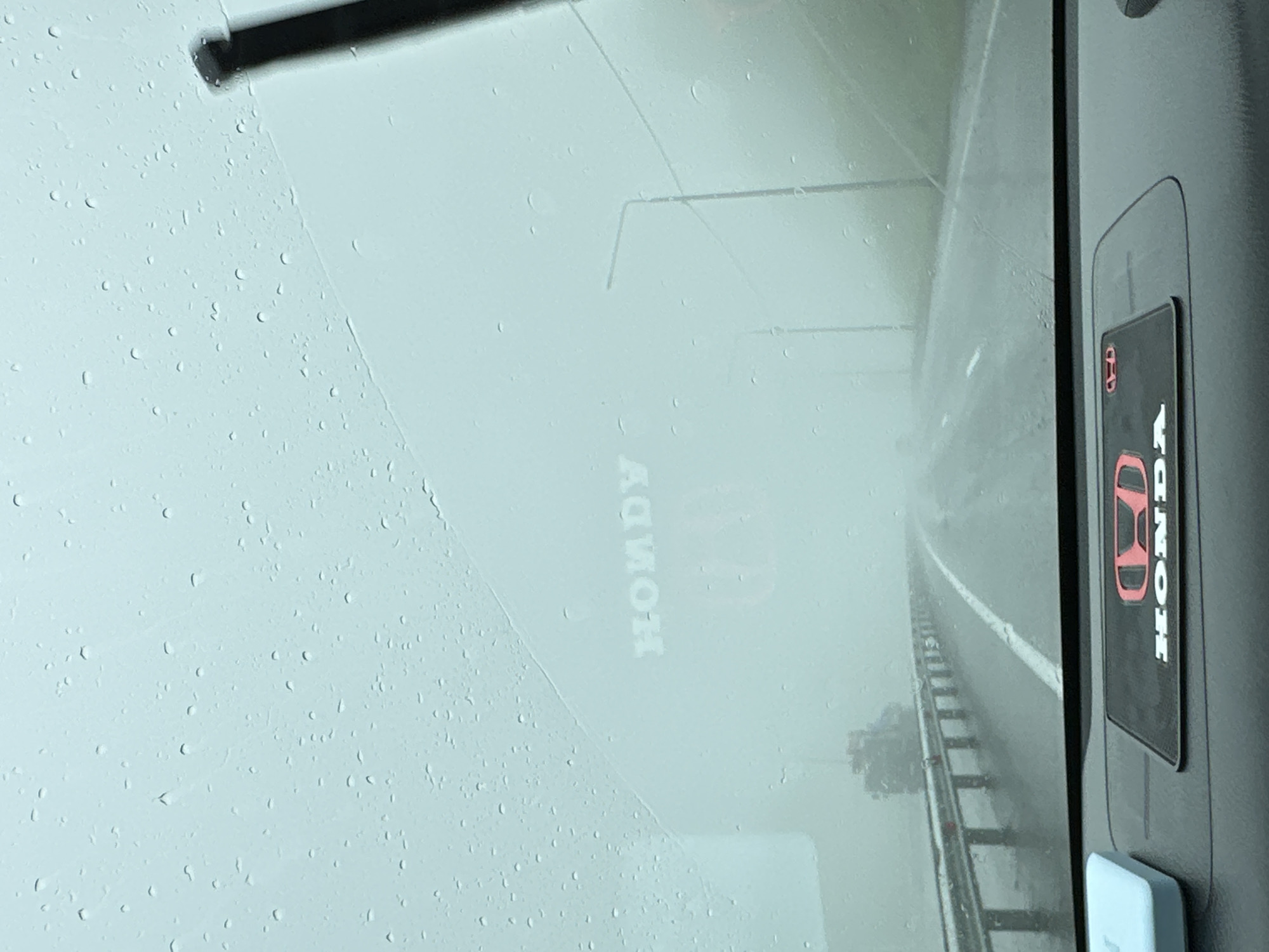 A car's windshield is covered in raindrops, obscuring the view of a foggy highway. In the distance, streetlights and a truck are barely visible. The dashboard features a Honda logo. The scene is the city of Svetly, Russia.