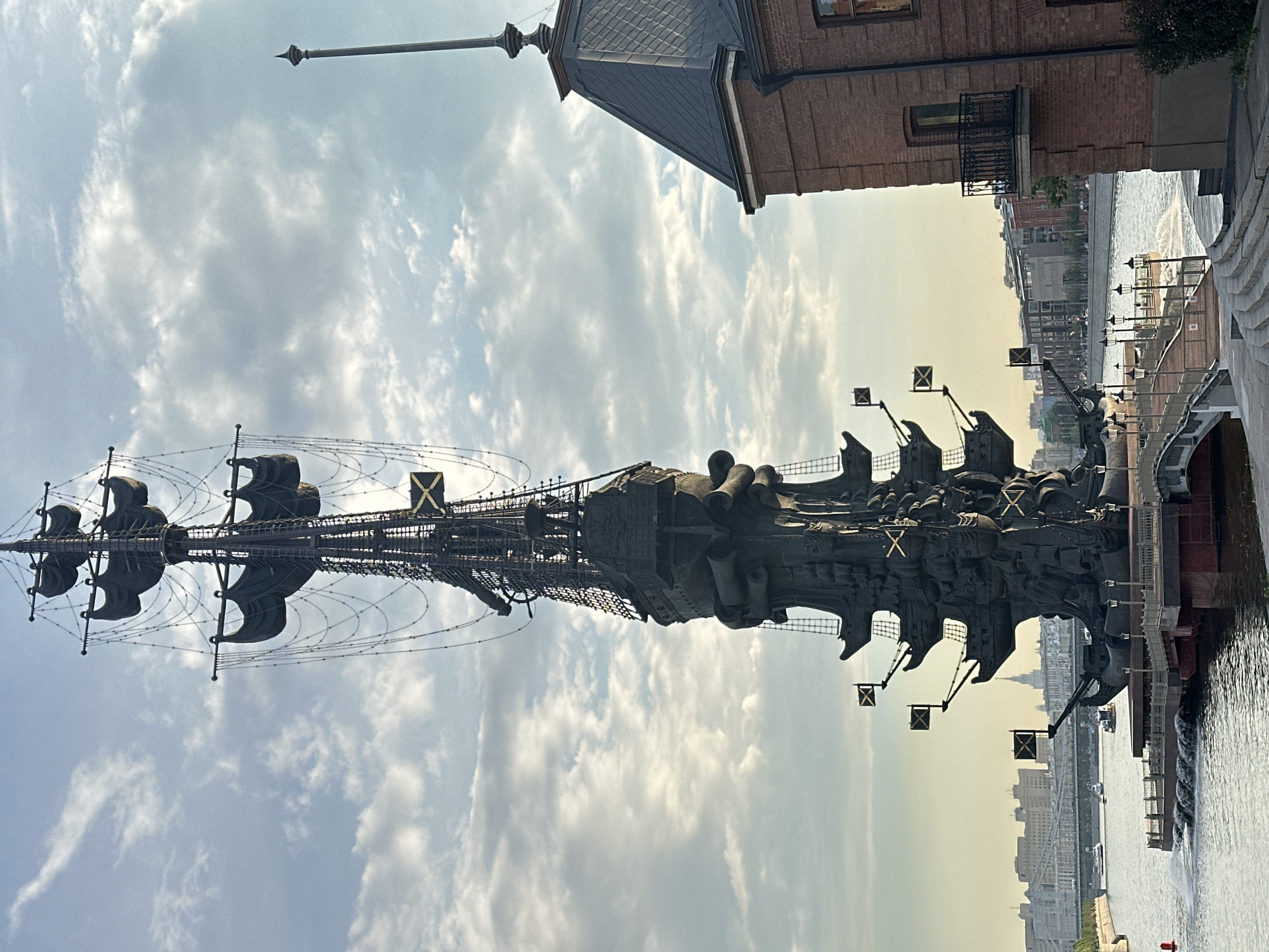 The image captures the Peter the Great Statue in Moscow, Russia, situated on an artificial island in the Moskva River. The colossal bronze sculpture depicts Peter the Great standing on the prow of a ship, towering over the water. The statue is intricately detailed, featuring multiple ship decks, masts with tattered sails, rigging, cannons, and flags. The background reveals the Moscow cityscape, with buildings and bridges visible along the riverbanks. The sky above is partly cloudy, with sunlight filtering through. To the right of the statue, a red brick building with a dark, pitched roof and a small balcony is partially visible, its architectural style contrasting with the monument. The water of the Moskva River reflects the sky and the surrounding structures, with some light ripples on its surface. The overall scene suggests a daytime view of this prominent landmark.