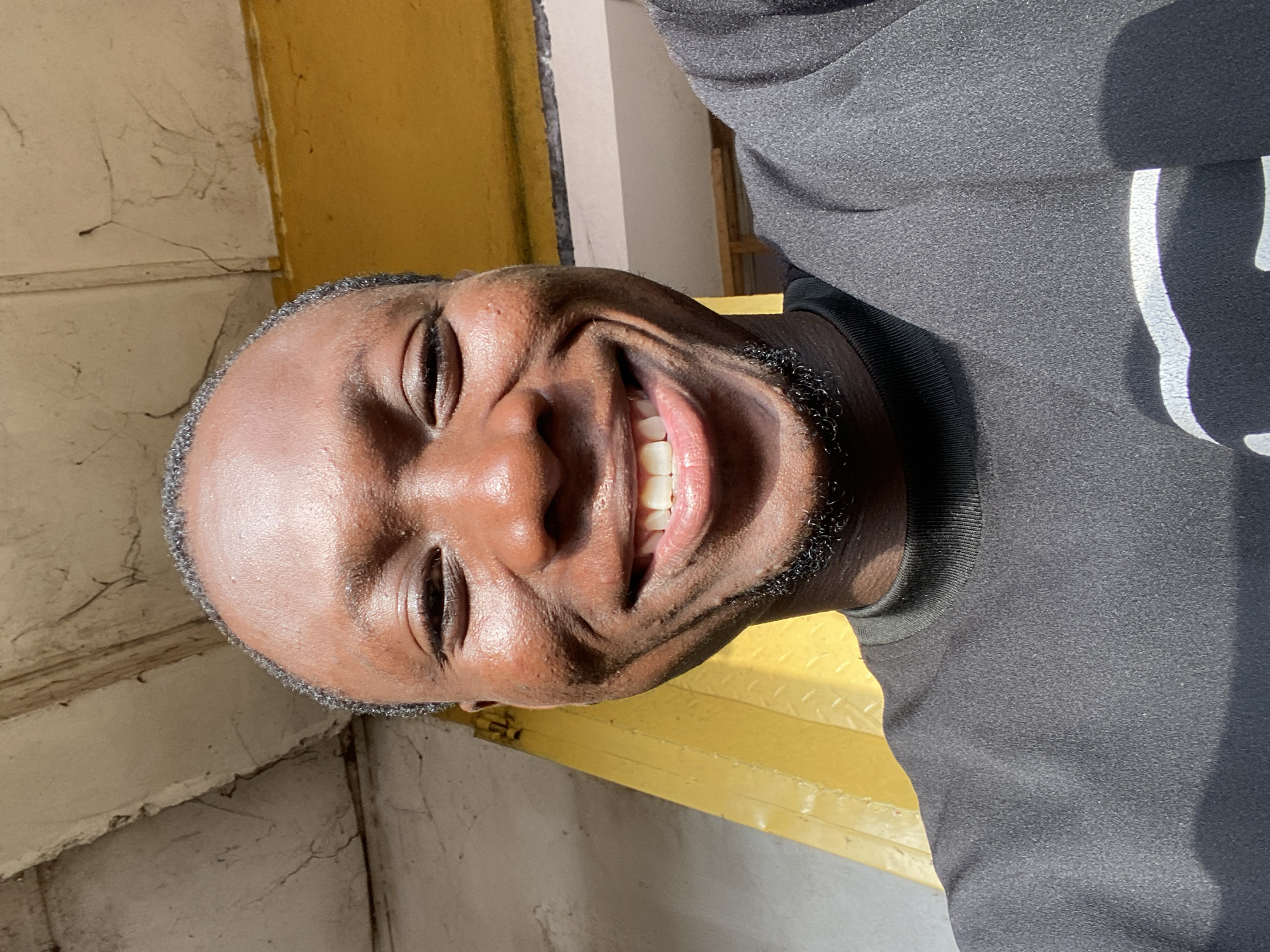A dark-skinned male with short hair and a goatee is pictured in a close-up, head-and-shoulders selfie. He is smiling widely, showing his teeth. He is wearing a black long-sleeved shirt with a high neckline. The background consists of a yellow wall and a white, textured overhang with some cracks. The lighting suggests an outdoor environment during the day. The overall scene is casual and appears to be taken in an urban setting. The provided information states this is in Bukuru, Nigeria.