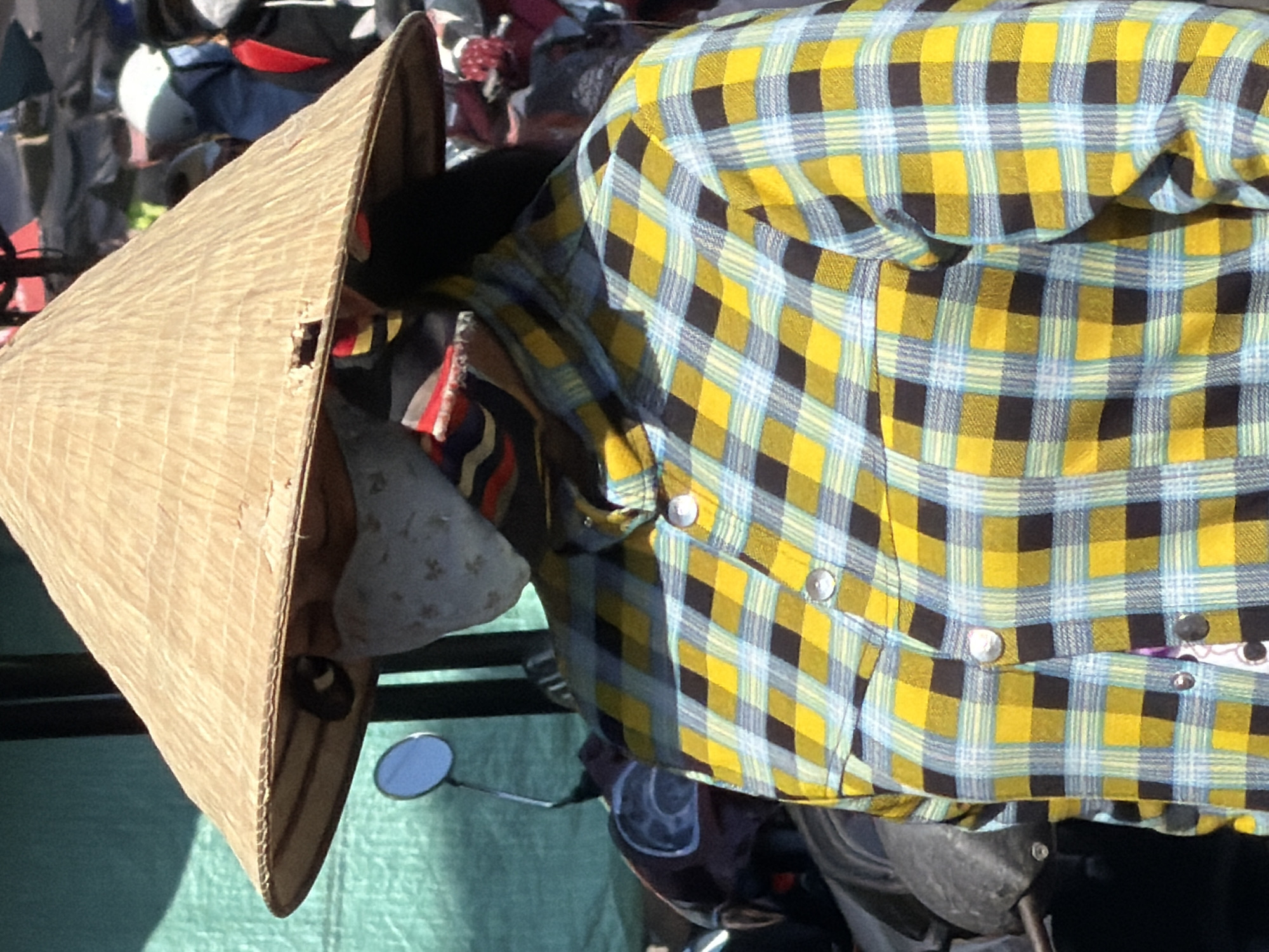 A person wearing a conical hat and a plaid shirt stands in front of a stall in Nha Trang, Vietnam. The hat is made of straw and has a hole in the brim. The person is also wearing a mask that covers their face. The shirt is yellow and blue plaid. In the background, there are motorcycles and other people. The scene appears to be a marketplace.