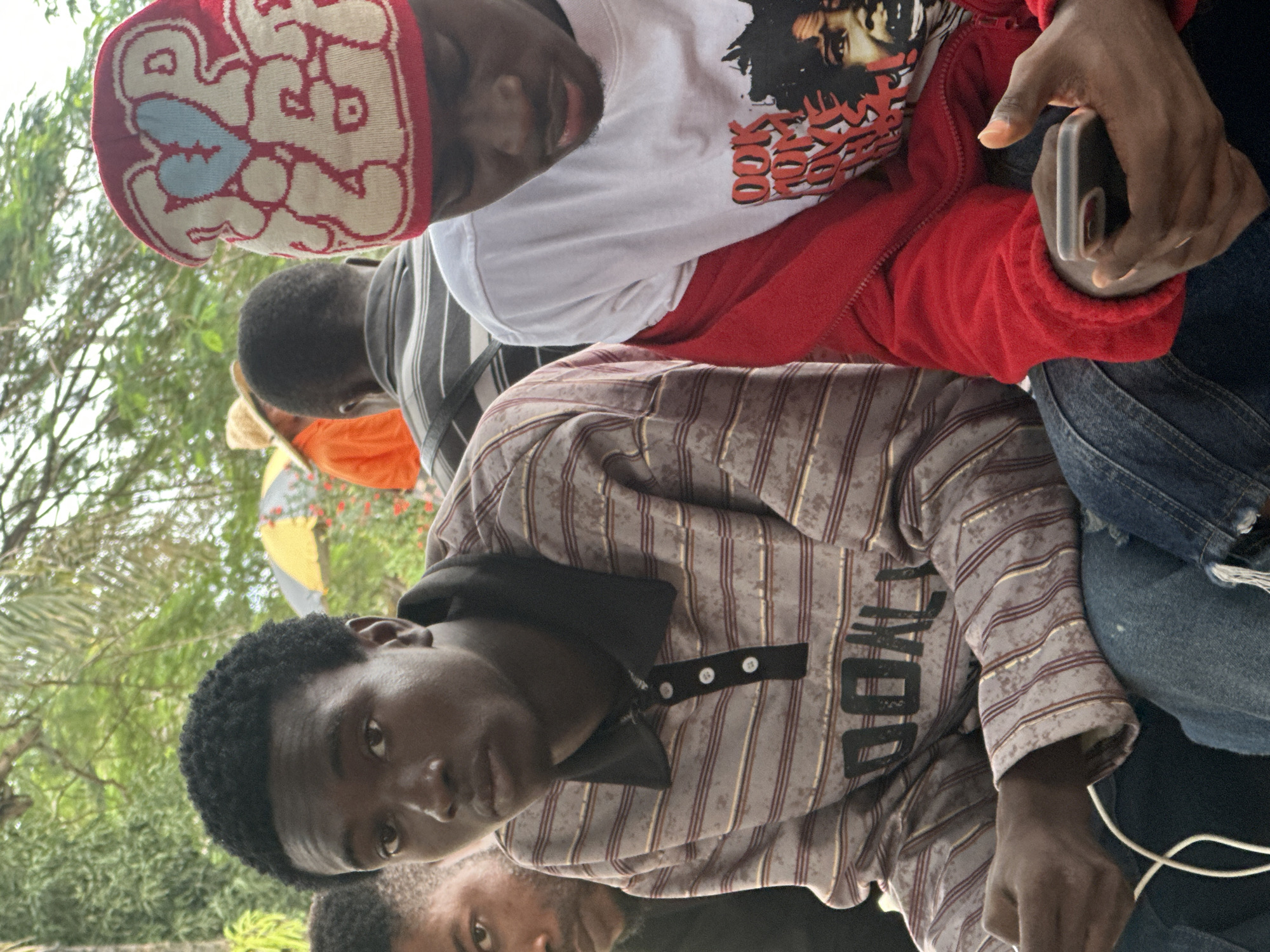 In Ihiagwa, Nigeria, three young men are visible in an outdoor setting with trees and foliage in the background. The man on the left is looking directly at the camera. He has dark, curly hair and is wearing a striped polo shirt with a black collar and white buttons. A white cable, possibly for headphones or charging, is visible in his lap. To his right, another young man is looking down at his phone, which he is holding in his right hand. He has dark skin and is wearing a white t-shirt with a red zippered jacket over it. The t-shirt has a graphic print on the front featuring a face and text. He is also wearing a red beanie with a cream-colored pattern including a blue heart. Behind these two men, another young man in a striped shirt and a person in an orange shirt and straw hat are partially visible. The overall atmosphere appears casual and relaxed.