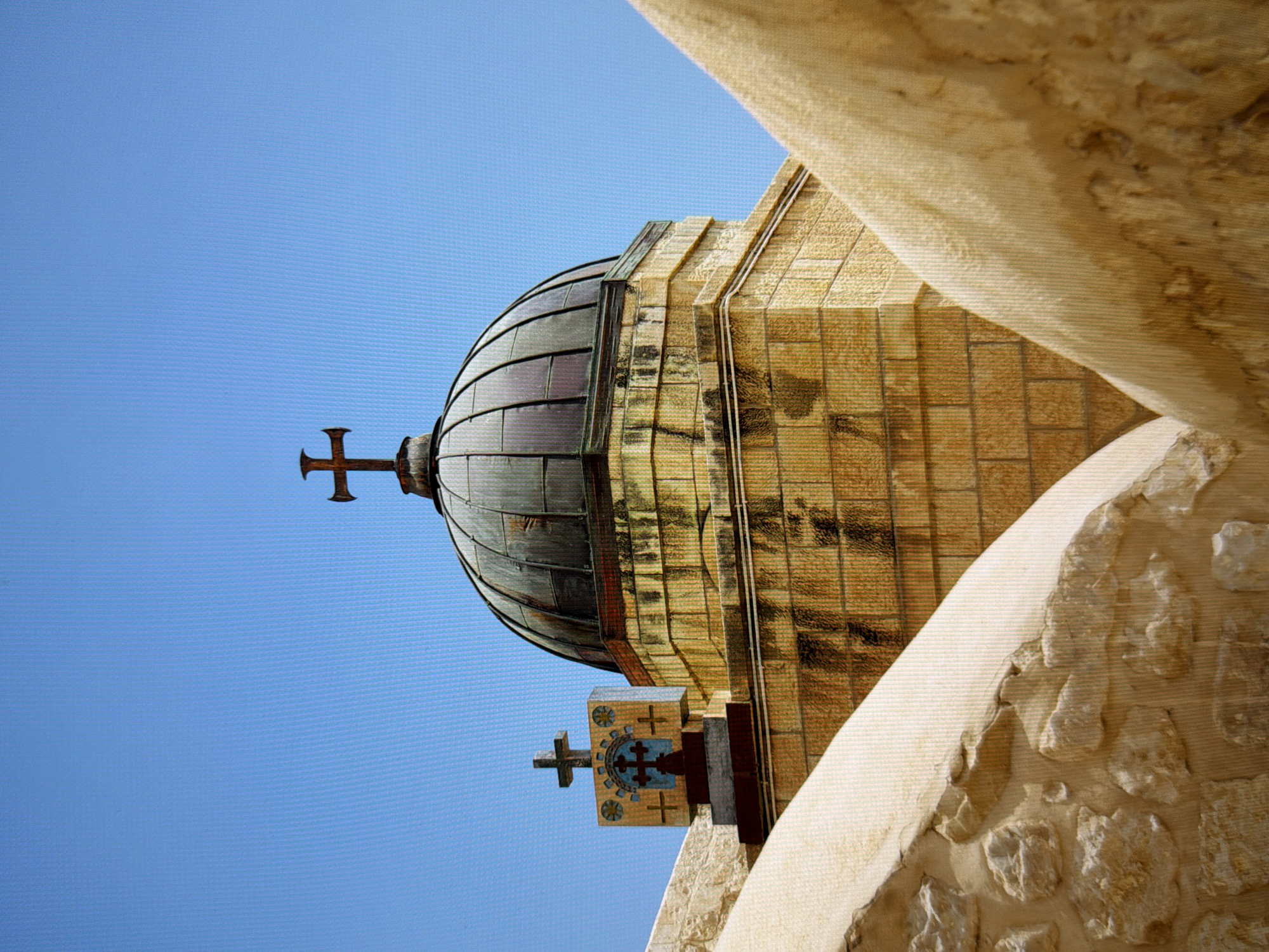 The image shows a close-up of a dome atop a stone building. The dome is dark, possibly made of metal, and appears aged. It is topped with a large cross. Below the dome, the building is constructed of rough-hewn, yellowish stone blocks. To the left of the main dome, a smaller structure with a cross on top is visible, adorned with decorative elements. In the foreground, curved, light-colored stone walls partially obscure the view of the building. The sky is a clear, bright blue, suggesting a sunny day. The architectural style suggests a religious building, possibly a church or monastery. This image does not provide any information about the city of Kyiv, Ukraine, or any specific location within it.