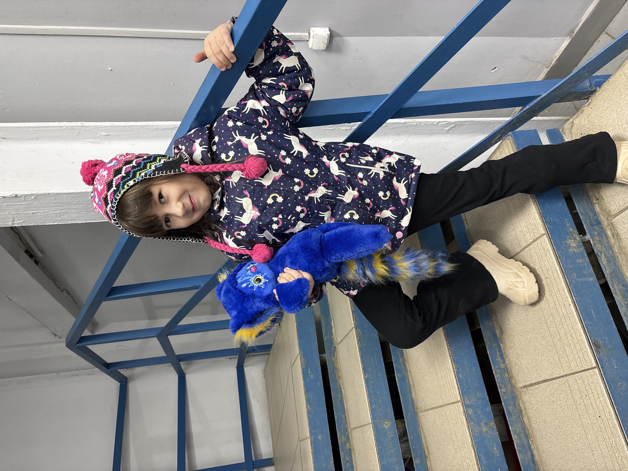 A young female child, approximately 5-8 years old, with dark brown hair and bangs, is positioned on a step of an indoor utilitarian stairwell. She wears a multicolored knitted winter hat with a prominent pink pom-pom and braided ear flaps, a navy blue hooded jacket patterned with white unicorns and pink rainbows/stars, black full-length pants, and light beige chunky-soled shoes. Her right hand grips a blue painted metal handrail. With her left arm, she holds a bright blue plush toy, approximately 30-40 cm in length, which features distinctive yellow fur on its ears and a striped tail. The toy has a stylized face with white and blue markings around its eyes and a pink nose. The child looks directly at the camera with a slight smile.
The stairwell steps are covered with light beige square tiles, showing minor wear and dirt accumulation, and the risers are painted blue. The walls are painted in two distinct tones: an off-white or light gray upper section and a darker gray lower section. A white electrical outlet or switch is mounted on the darker gray wall to the child's right. Visible ceiling infrastructure, including a white painted beam and pipes, is present in the upper left background. The scene is consistent with a generic indoor stairwell, and no specific landmarks or architectural features are identifiable to pinpoint a precise location within Izhevsk, Russia.