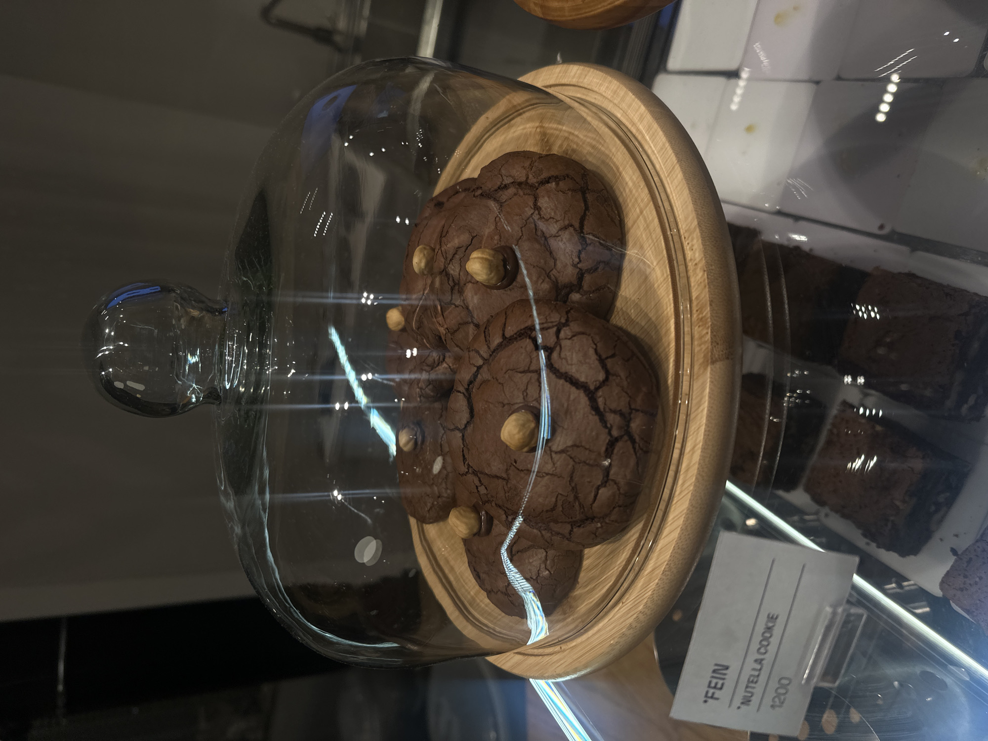 A clear glass dome sits atop a wooden tray displaying several dark brown cookies studded with nuts, possibly hazelnuts, inside a pastry display case in Yerevan, Armenia. The tray is light-colored wood, contrasting with the cookies' rich, dark hue. The pastry case also contains dark brownies. A price tag is visible, reading "*FEIN Nutella Cookie 1200" and presumably indicating the price of the cookie is 1200 Armenian Drams. The pastry case is partially reflective, blurring the background. The scene is indoors, likely a bakery or café in Yerevan, Armenia.