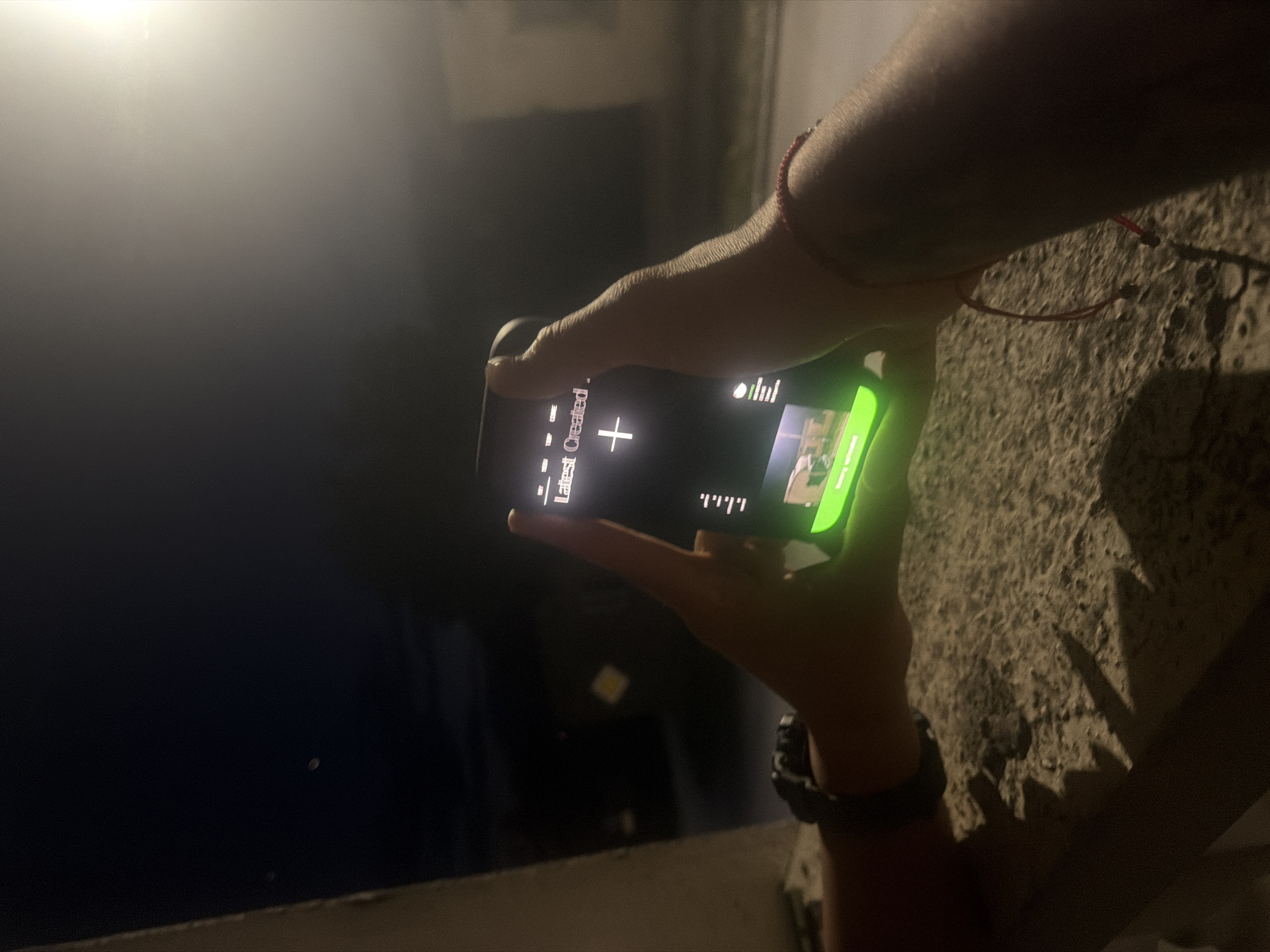 The image captures a close-up of a person's hands holding a smartphone in a landscape orientation, likely outdoors at night or in a very dimly lit indoor environment. The hands are positioned in the lower-right portion of the frame, with the illuminated phone screen as a central focus.
The subject's forearms and hands are visible. A thin red string bracelet is worn on the left wrist, while the right wrist features a dark-colored watch. The skin tone appears moderately tan.
The smartphone screen displays a user interface with several elements. On the left, the text "Latest Created" is clearly visible, followed by a large white plus sign ("+") icon. To the right of these, a series of short, vertical white lines, resembling a volume indicator or sound waves, are displayed. A small, indistinct image thumbnail can be seen further to the right. The bottom edge of the phone screen is accented with a bright, vibrant green highlight or border.
To the right of the person's hands, a rough, textured concrete surface, possibly a wall or a balcony railing, is partially visible and dimly lit, casting subtle shadows. The background beyond the phone is predominantly dark, indicating a nighttime setting, with only faint, indistinct light sources or shapes in the distance. The overall mood is quiet and focused on the interaction with the digital device.