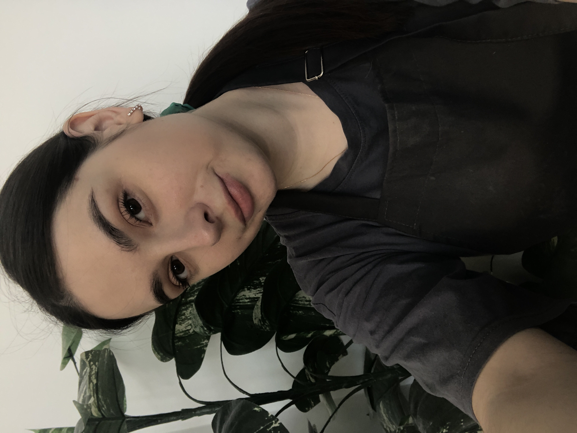A selfie of a fair-skinned woman with dark hair and eyebrows. She has brown eyes and wears delicate earrings. She is wearing a dark gray t-shirt and a black apron. A thin gold necklace is visible. In the lower portion of the frame, there is a potted plant with dark green and white leaves. The background is a plain white wall. The image does not contain sufficient information to determine the location is Chaikovsky, Russia.