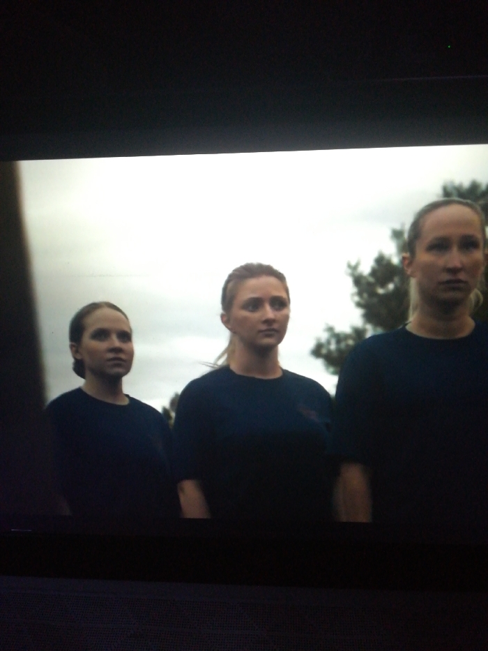 Three Caucasian women wearing matching navy t-shirts stand in a row. They all face forward with serious expressions. The woman in the middle has her blonde hair pulled back into a ponytail. The woman on the right has her hair pulled back as well, revealing her forehead. The woman on the left has her blonde hair pinned back, away from her face. A cloudy sky and trees are visible in the background.
