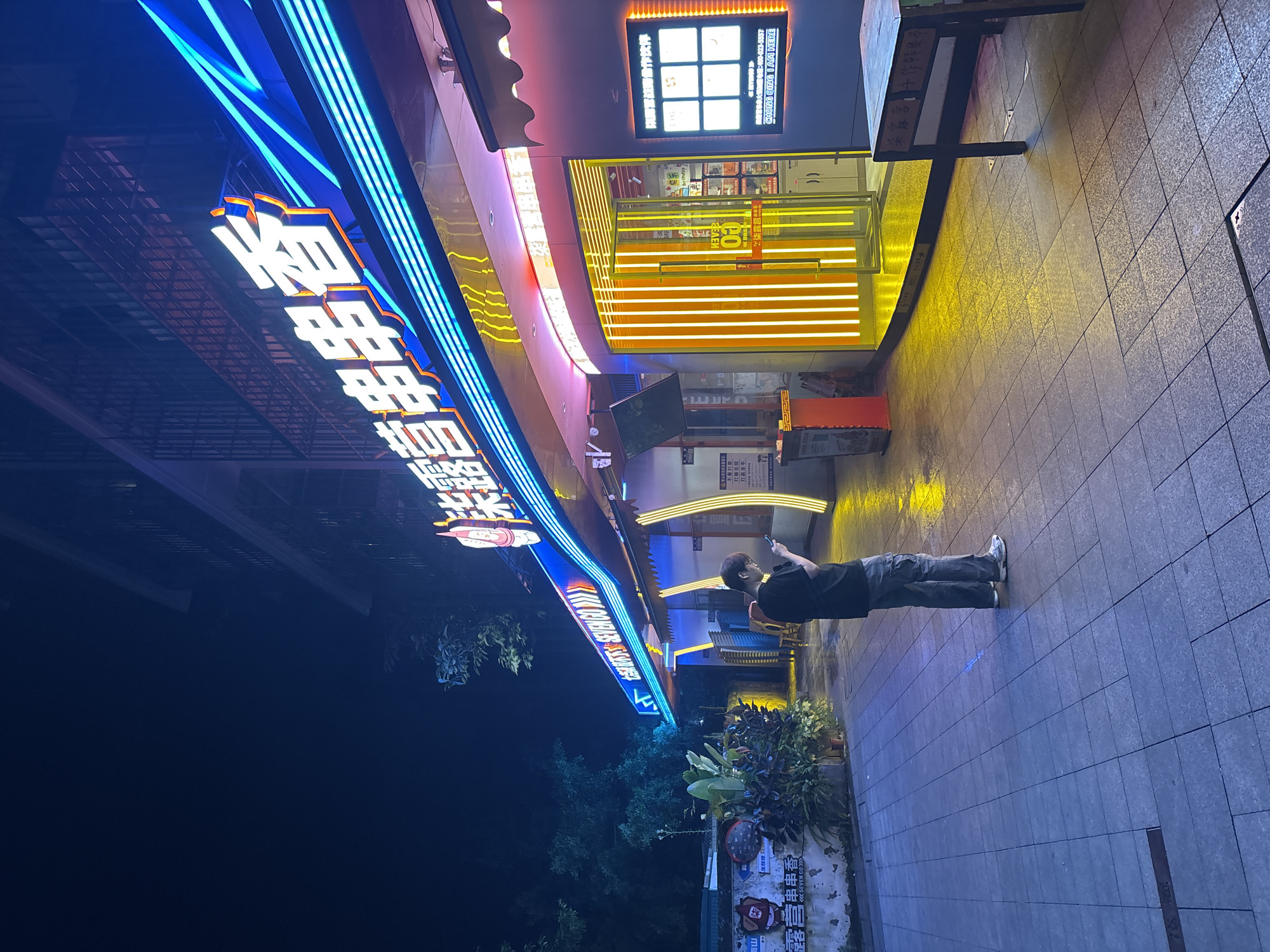 At night in Nan'an District, China, a person in dark clothing stands on a tiled sidewalk, looking up at a brightly lit building facade. The building features vibrant blue and orange neon signs with Chinese characters, specifically "露营串串香" (Lùyíng chuàn chuàn xiāng), indicating a restaurant specializing in skewers. The adjacent storefront has orange horizontal light bars, while a menu display is visible at the top. The sidewalk is illuminated by yellow and blue light reflecting from the signs. Adjacent to the sidewalk, there are some plants and signboards visible with Chinese characters on them.