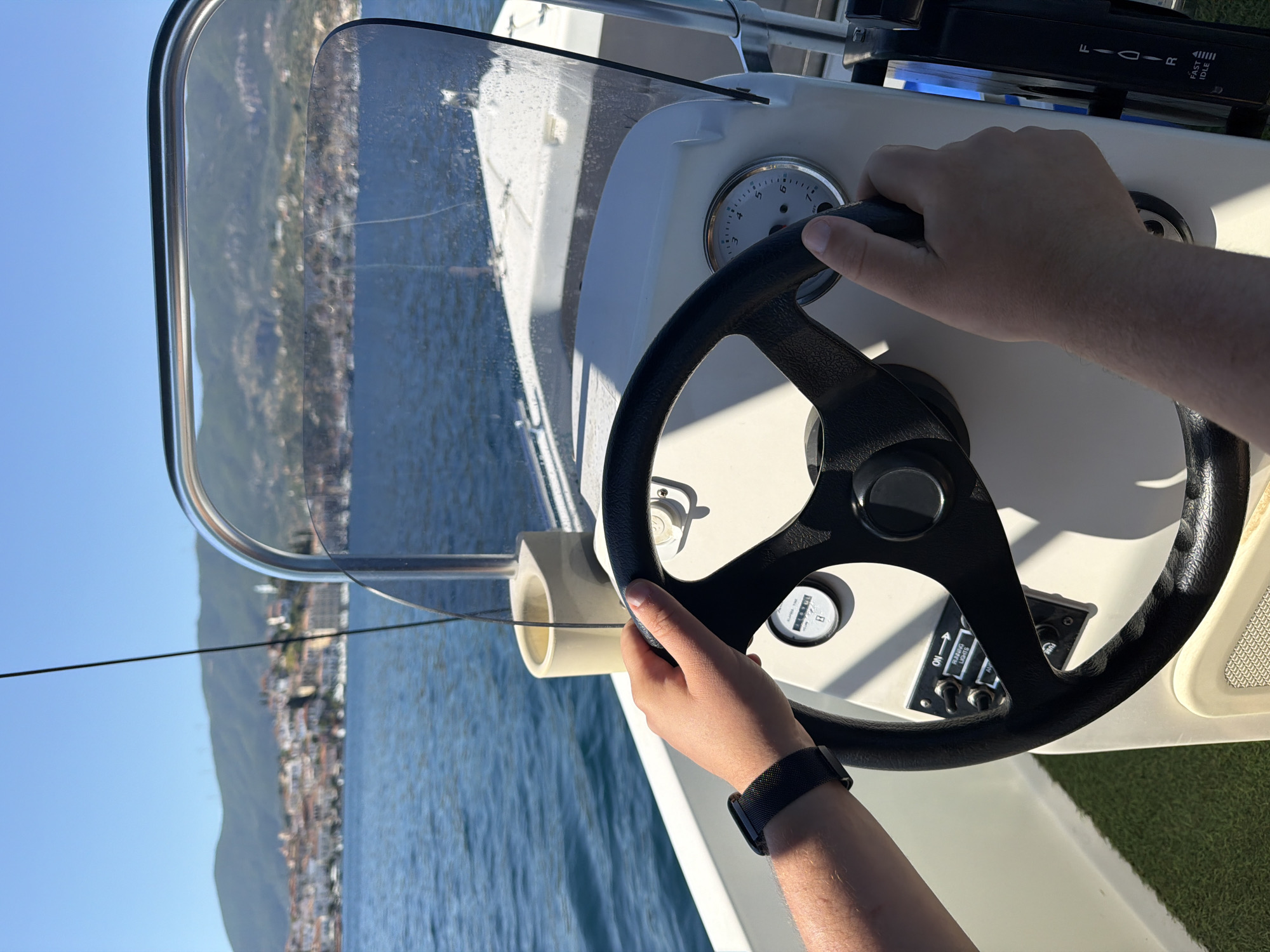 The image provides a first-person perspective from the helm of a boat, with a person's hands actively gripping a black steering wheel. The left hand, wearing a dark mesh-band watch, is positioned on the lower left of the wheel, while the right hand rests on the upper right.
The boat's white console features several instruments. A circular gauge, likely a tachometer, displays numbers from 1 to 8, with its needle pointing between 2 and 3. To its left, a small rectangular gauge shows numerical readings "101.6", "101.5", and "101.7", possibly an hour meter. Below the steering wheel, a panel with switches is visible, labeled "ON", "BOW LIGHTS", and "PUMP". Above the console, a black throttle or gear lever shows positions "F" (Forward), "I", "D" (Drive), "R" (Reverse), and "FAST IDLE".
A clear, curved windshield, slightly smudged, offers a view of the outside. Beyond the boat, a vast body of deep blue water, likely the sea, stretches out under a bright, clear blue sky. In the distance, a mountainous coastline with visible buildings of a coastal town can be seen, consistent with the location of Poros, Greece. The scene is illuminated by strong daylight, casting shadows on the dashboard and the person's hands, indicating a sunny day. The overall impression is one of calm navigation on the water.