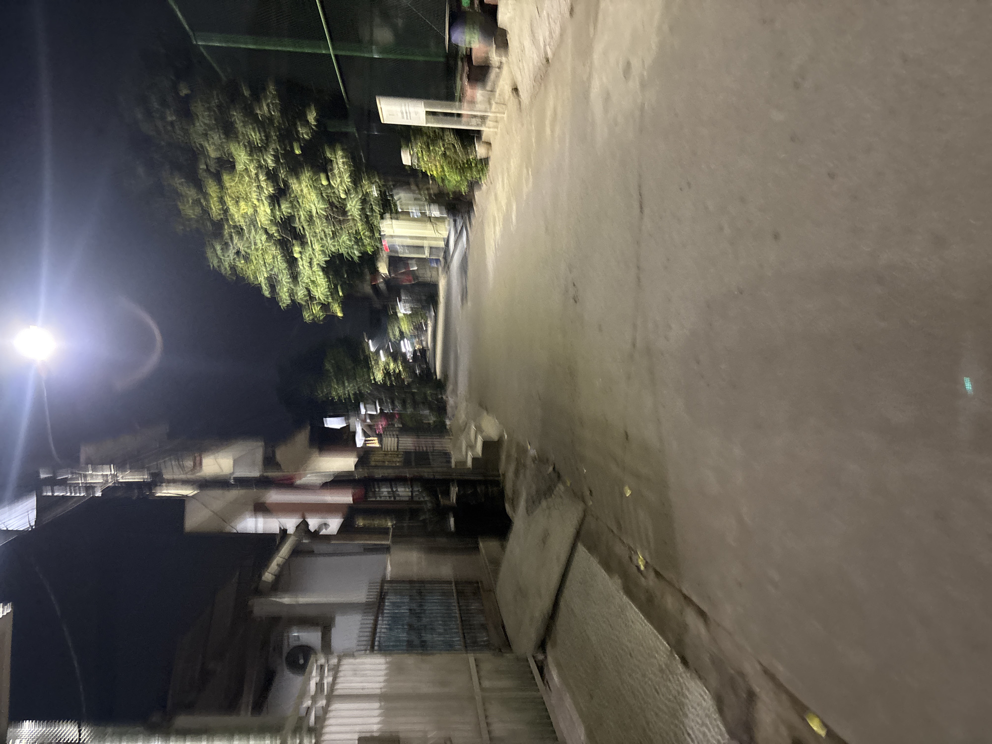 The image depicts a street scene at night in Telagamurni, Indonesia. A street lamp illuminates the paved road, casting long shadows. Buildings line the left side of the street, featuring a white facade, a metal gate, and an air conditioning unit. On the right, lush green trees with dense foliage are visible, bordering a fenced area, possibly a sports court. Various objects like trash cans, pots with plants, and a signpost are scattered along the roadside. The overall impression is that of a quiet residential or commercial area illuminated by artificial light.