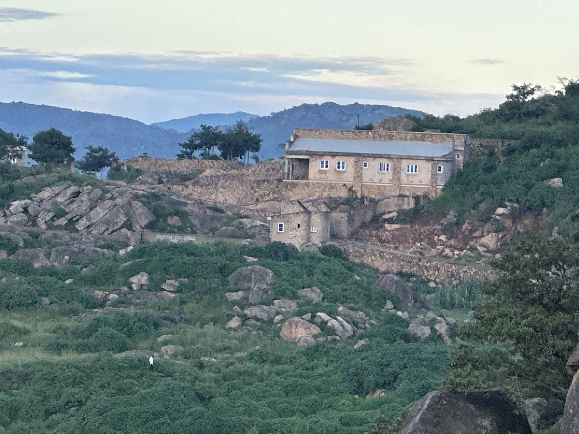 The image depicts a landscape scene with a prominent building situated on a rocky, elevated terrain. The building appears to be constructed of stone and has a light-colored facade with multiple windows. The roof is light gray and appears to be made of metal. The surrounding environment is characterized by rocky outcrops, dense green vegetation, and rolling hills in the background under a cloudy sky. In the foreground, a lone individual, possibly a man in a white shirt and dark pants, is visible walking through the vegetation. The architectural style and the natural landscape suggest a location in a hilly or mountainous region. This scene is consistent with the geography and architecture found in parts of Jos, Nigeria, known for its plateau landscape and stone structures.
