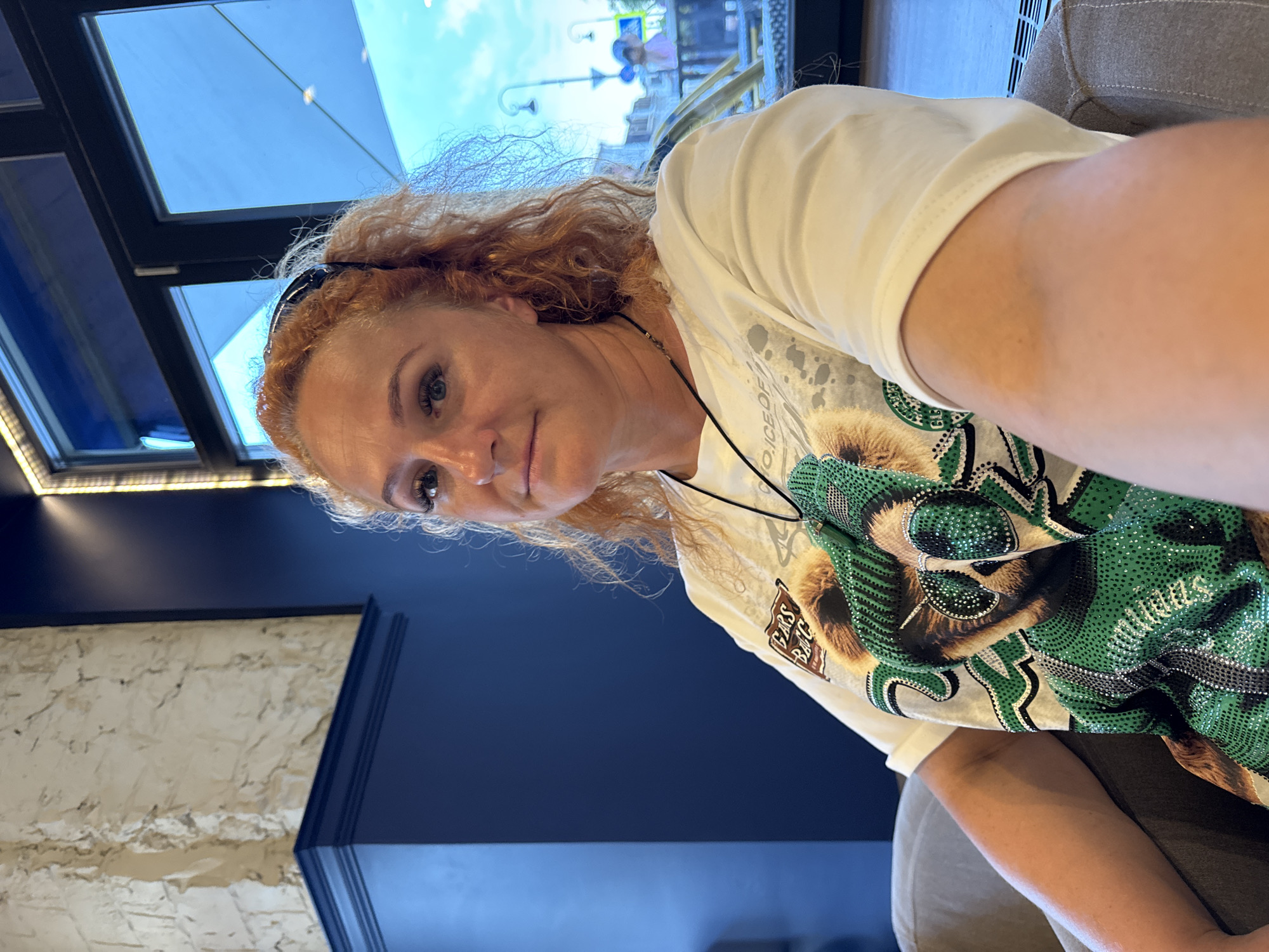 A mid-shot, selfie-style photograph features a woman with long, curly red hair, wearing a white t-shirt with a graphic of a bear wearing sunglasses and a sparkly green outfit. She is seated in a chair, looking towards the camera. Behind her, a large window reveals a view of a street scene in Kazan, Russia, with buildings, lampposts, and people walking by. The interior of the room includes a dark blue wall and a textured, light-colored wall with a dark blue trim. The lighting suggests it is daytime.