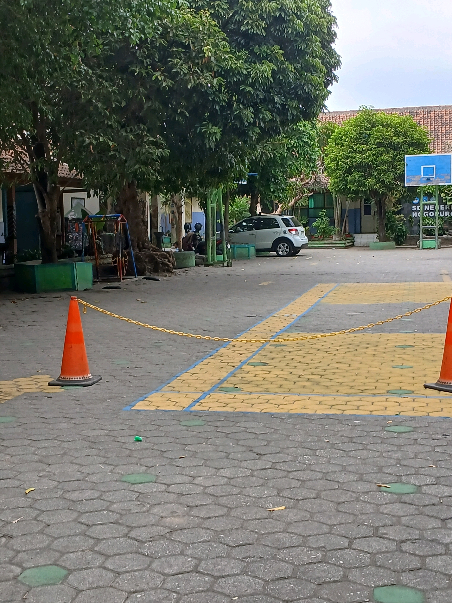 The image captures an outdoor scene at an educational facility, likely a school, in Depok, Indonesia. In the foreground, a grey hexagonal paver ground is visible, with several green rectangular inlays. An orange traffic cone with a yellow chain attached to its top sits on the pavers. A chain extends from the cone diagonally across the image towards the right. Behind the cone, a section of the ground is marked with yellow and blue rectangles, suggesting a court or designated area.
Further in the background, a white SUV is parked. Trees with dense green foliage are prominent on the left and in the middle of the image. A playground structure with red and blue elements is partially visible behind a large tree trunk on the left. Buildings with tiled roofs and some windows are present. A blue basketball hoop is mounted on a structure on the right side of the image, with text on a white sign below it that appears to read "SD NEGERI" followed by a partial inscription of a location name. Two individuals, dressed in dark clothing, are seated or standing in the shade of a tree near a building on the right. The sky is overcast. The overall environment suggests a schoolyard.