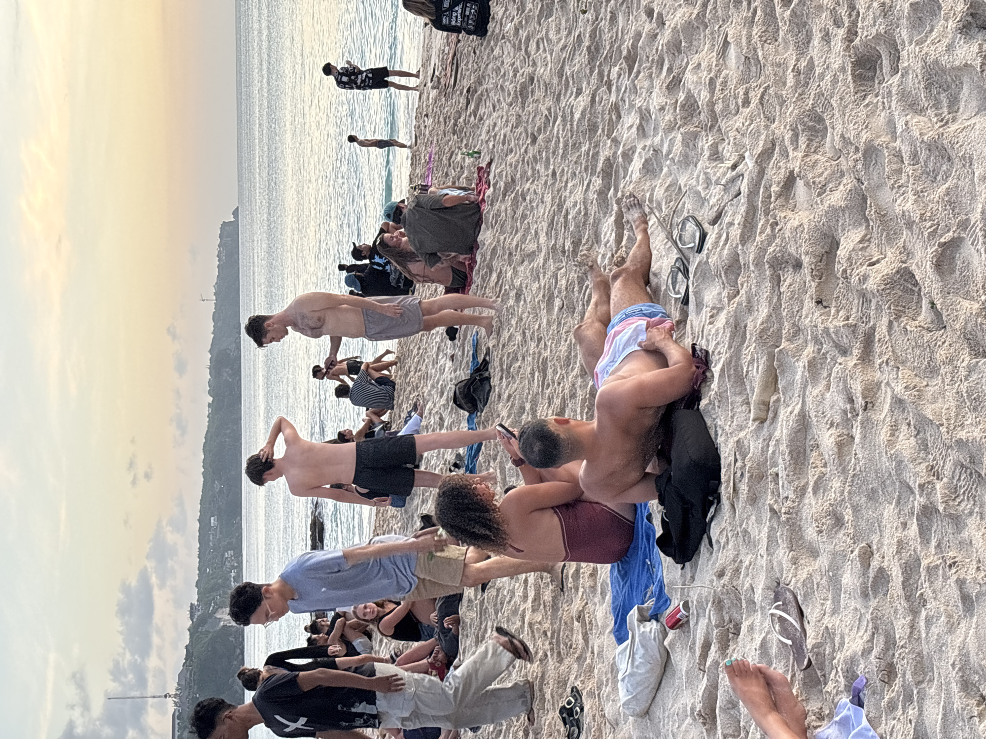 The image depicts a crowded beach scene in Kuta Selatan, Indonesia. A group of people are gathered near the water's edge, with several individuals standing, walking, or wading in the sea. Closer to the foreground, people are sitting or lying on the sand. Some individuals are using mobile phones. Beach accessories, such as bags, towels, and flip-flops, are scattered around. The background shows a coastline with some visible structures and vegetation. The sky is partly cloudy.