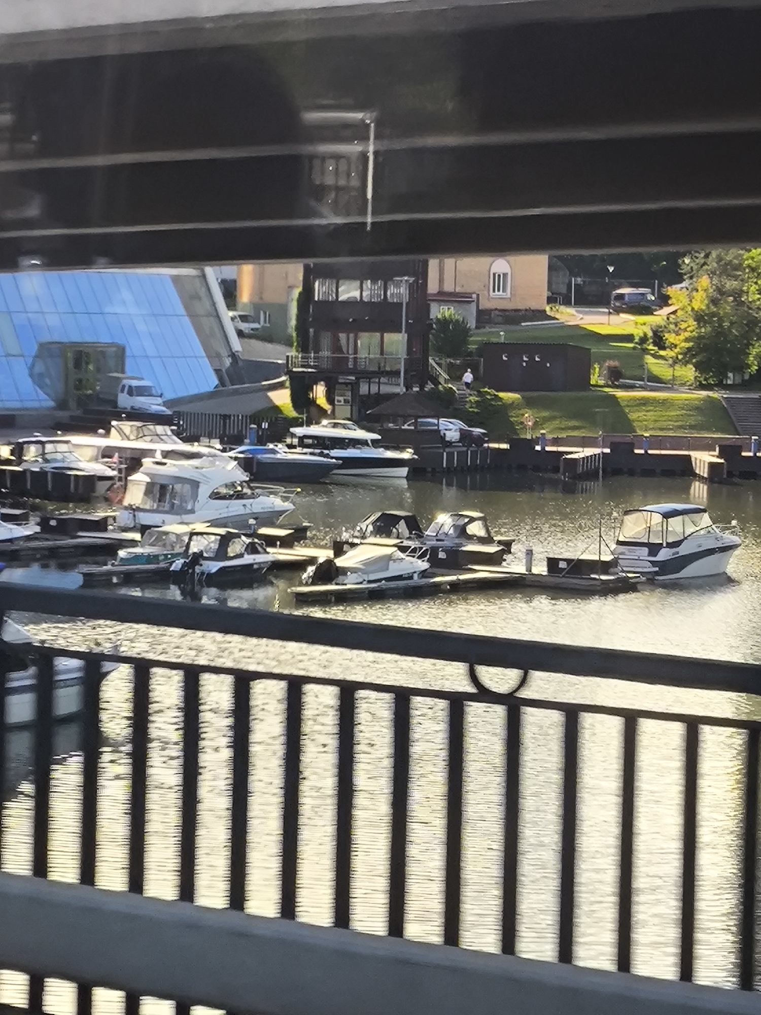 The image shows a view of a marina filled with several boats docked at wooden piers. The boats are of various sizes and types, including speedboats and larger cabin cruisers, some with canvas coverings. The water in the marina is calm and reflects the sunlight, creating a shimmering effect.
In the background, there are buildings and trees. To the left, a modern building with a large blue glass roof is visible. Further back, there are residential-looking buildings with windows and pitched roofs, and some darker, more commercial structures. A green, grassy embankment rises behind the marina, dotted with trees, some of which are vibrant green, suggesting it's likely summertime. A few cars are parked on a road behind the embankment, and a single person is visible walking on the grass.
The lighting suggests it is late afternoon or early evening, with the sun casting long shadows and creating a warm, golden glow on the water and the landscape. The sky is not directly visible, but the quality of light indicates clear or partly cloudy weather. The scene appears peaceful and serene, typical of a calm day at a harbor. There is no visible text in the image.
