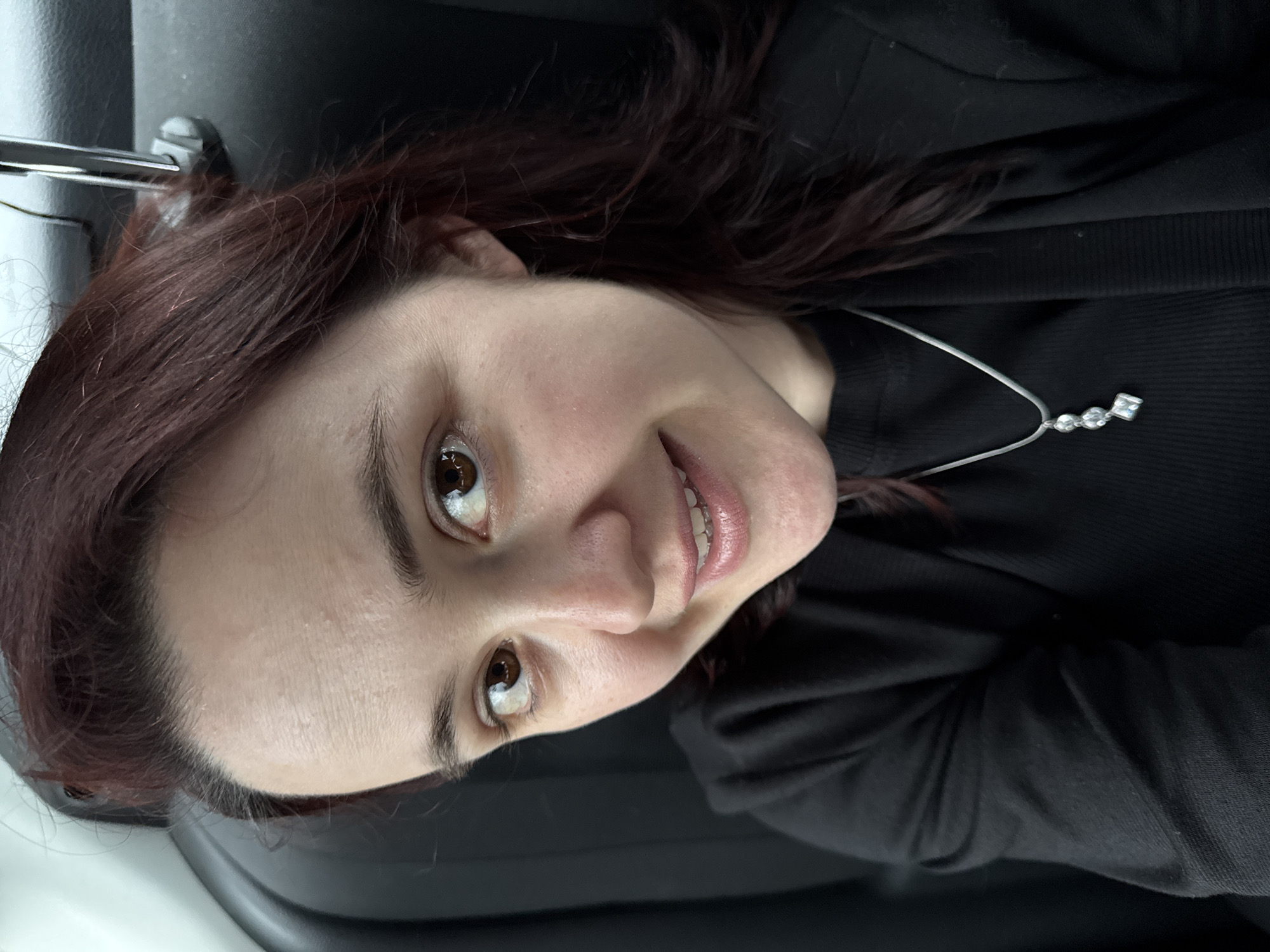 A woman with brown hair and light skin is photographed from an unusual, angled perspective. She is wearing a black jacket or sweater and a silver necklace with three small, clear stones or beads and a larger square stone at the bottom. The background is dark, appearing to be the interior of a vehicle, possibly a car, with part of the headrest visible on the left. The location of Sochi, Russia, is not discernible from the visual information provided.