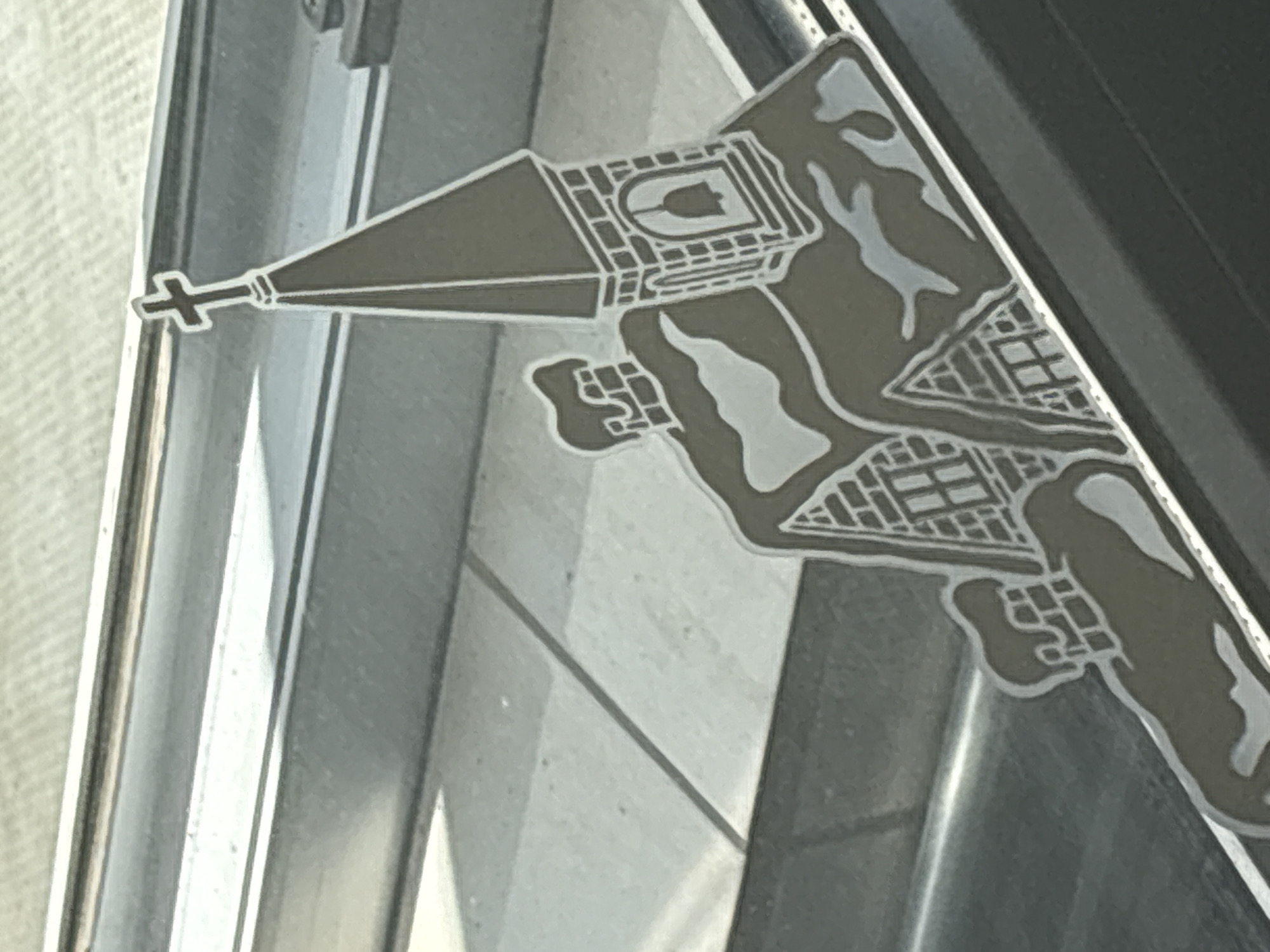 A close-up, angled shot shows a metallic decal of a snow-covered church. The decal features a steeple topped with a cross, a bell tower with a visible bell, and a building with brick detailing and chimneys. Snow is depicted on the roof and chimneys. The decal appears to be on a smooth, reflective surface, possibly a window or car door. The background is out of focus and appears to be an outdoor environment with tiled ground and horizontal metallic elements.