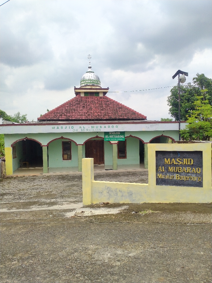 An exterior shot captures Masjid Al-Mubarooq, a mosque located in Kecamatan Kebakkramat, Indonesia, under an overcast sky. The mosque features light greenish-blue walls with darker green trim around its arched entranceways and windows. Its roof is covered with reddish-brown tiles, and a small, silver-colored dome topped with a finial sits centrally above the roofline. Two wooden double doors are visible at the main entrance.
In front of the mosque, a paved, somewhat rough ground leads up to the building. A low, yellow-painted wall or boundary runs across the foreground. Integrated into this wall on the right side is a larger, black rectangular sign with raised yellow text that reads: "MASJID AL MUBARAQ Mirukon Banjarharjo". Above the main entrance of the mosque, a banner displays "MASJID AL-MUBAROQ DK MIRUKUN". Additionally, a smaller, green hanging sign with "MASJID AL-MUBAROQ" and some indistinct smaller text is positioned centrally near the entrance.
To the right of the mosque, a pole with a speaker or light fixture is visible, alongside some green foliage from nearby trees. No people or specific activities are depicted in the static scene. The overall impression is a community mosque with functional and modest architecture, situated in what appears to be a non-urban setting during daytime.