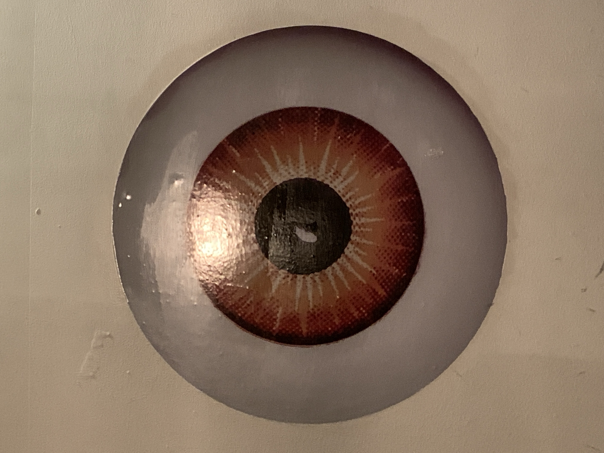 The image depicts a close-up of a stylized eye affixed to a light-colored surface, possibly a wall. The eye consists of a greyish-white sclera surrounding a textured, reddish-brown iris with radial markings. A black pupil is located in the center of the iris. There are reflections on the surface of the eye, suggesting it has a glossy finish. The background surface appears slightly textured. No people, specific objects other than the eye, actions, or interactions are visible. No location information can be discerned from the provided image. The prompt to include Barcelona, Spain, appears to be irrelevant.