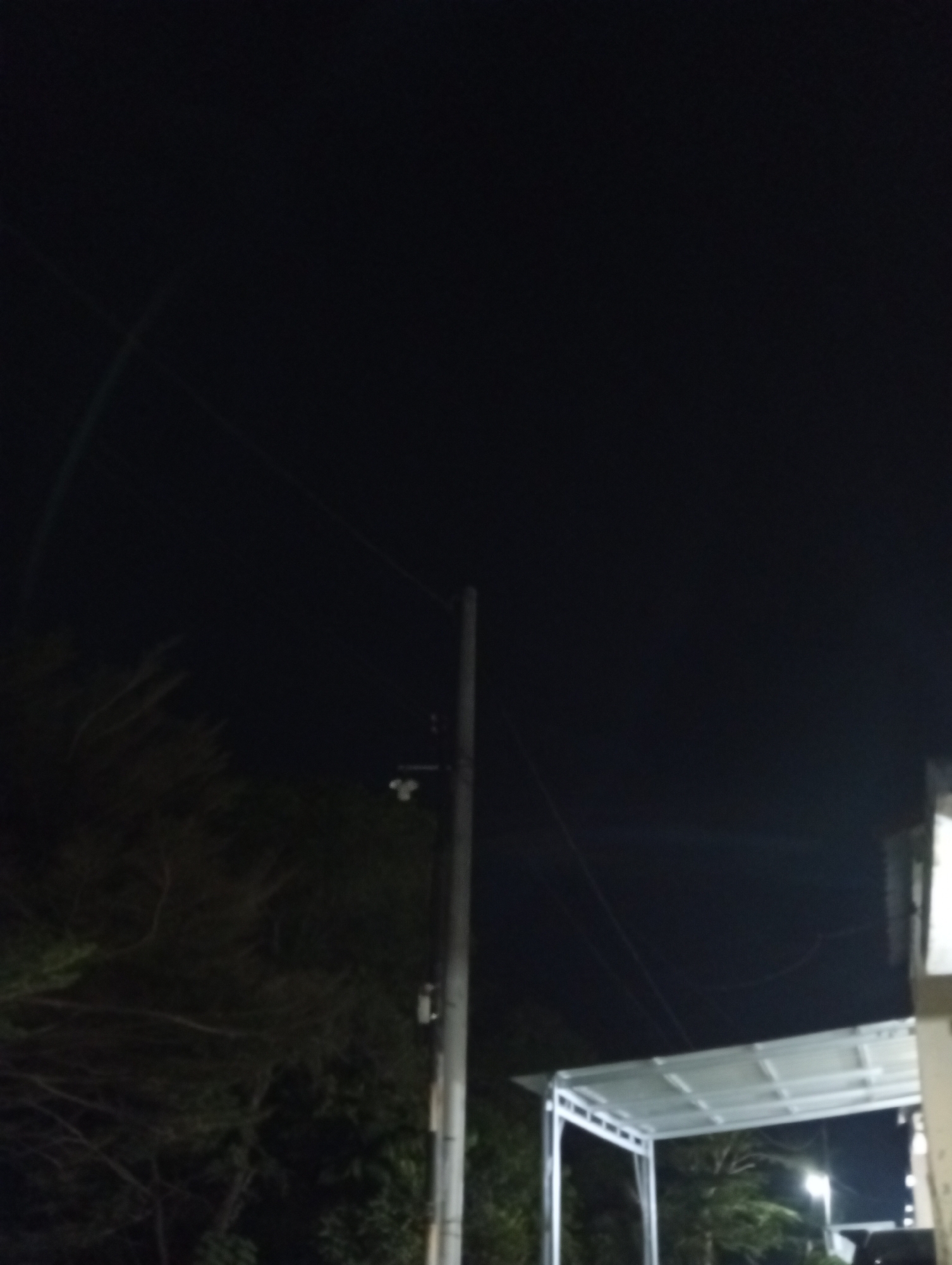 This is a nighttime, outdoor, low-light image showing a street scene with a utility pole, power lines, trees, and a structure that appears to be a carport or shelter. A small, white security camera is mounted on the utility pole. The overall environment is dark with minimal ambient light. The location is Kualu, Indonesia. There are no people or animals visible in the image.