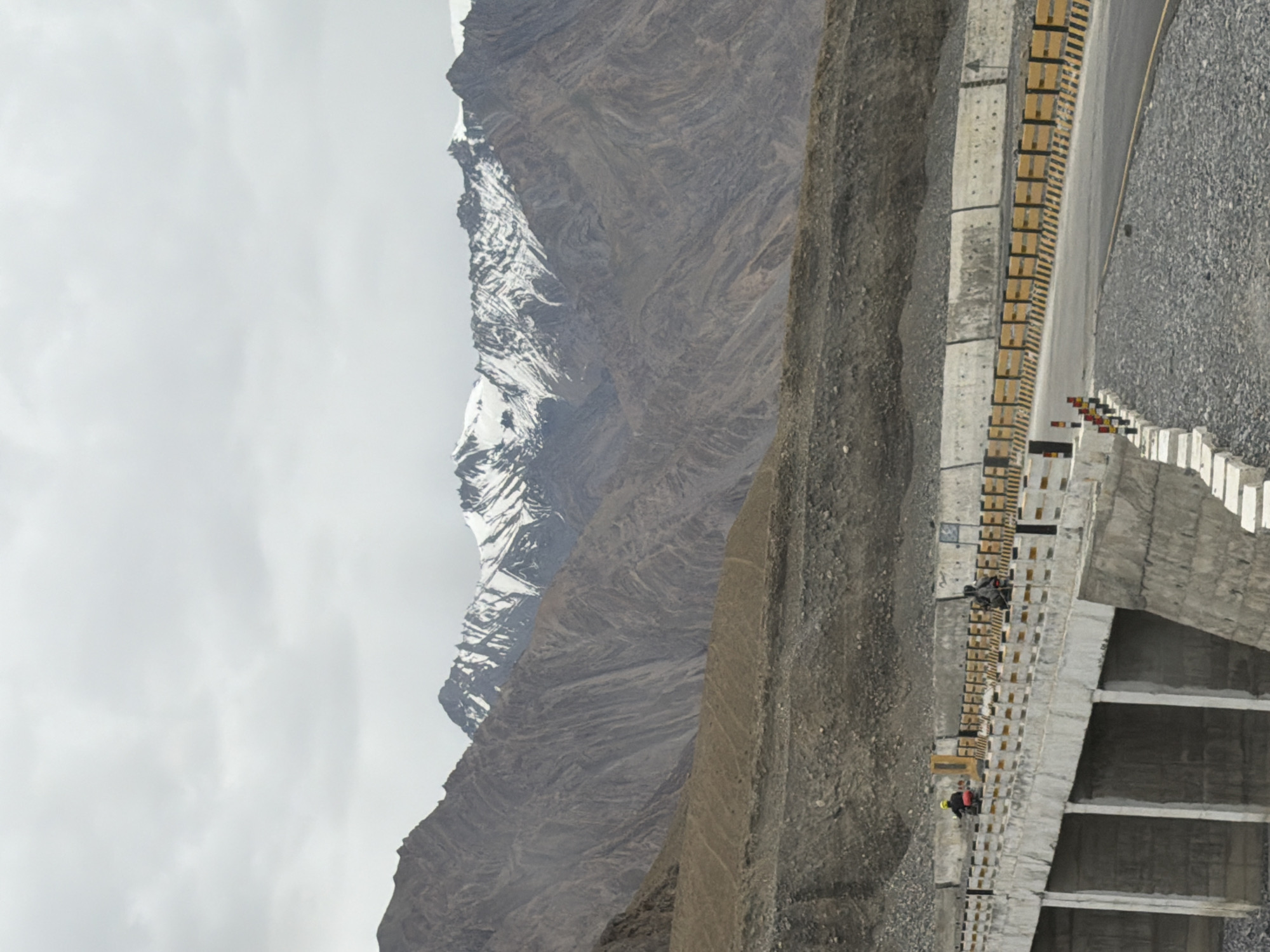 A motorcyclist wearing a yellow jacket and helmet rides on a paved road with a concrete bridge. The bridge has a yellow and black striped guardrail and concrete supports. In the background, a mountain range with snow-capped peaks is visible under a cloudy sky. The surrounding terrain is rocky and barren. The location is Zangla, India.