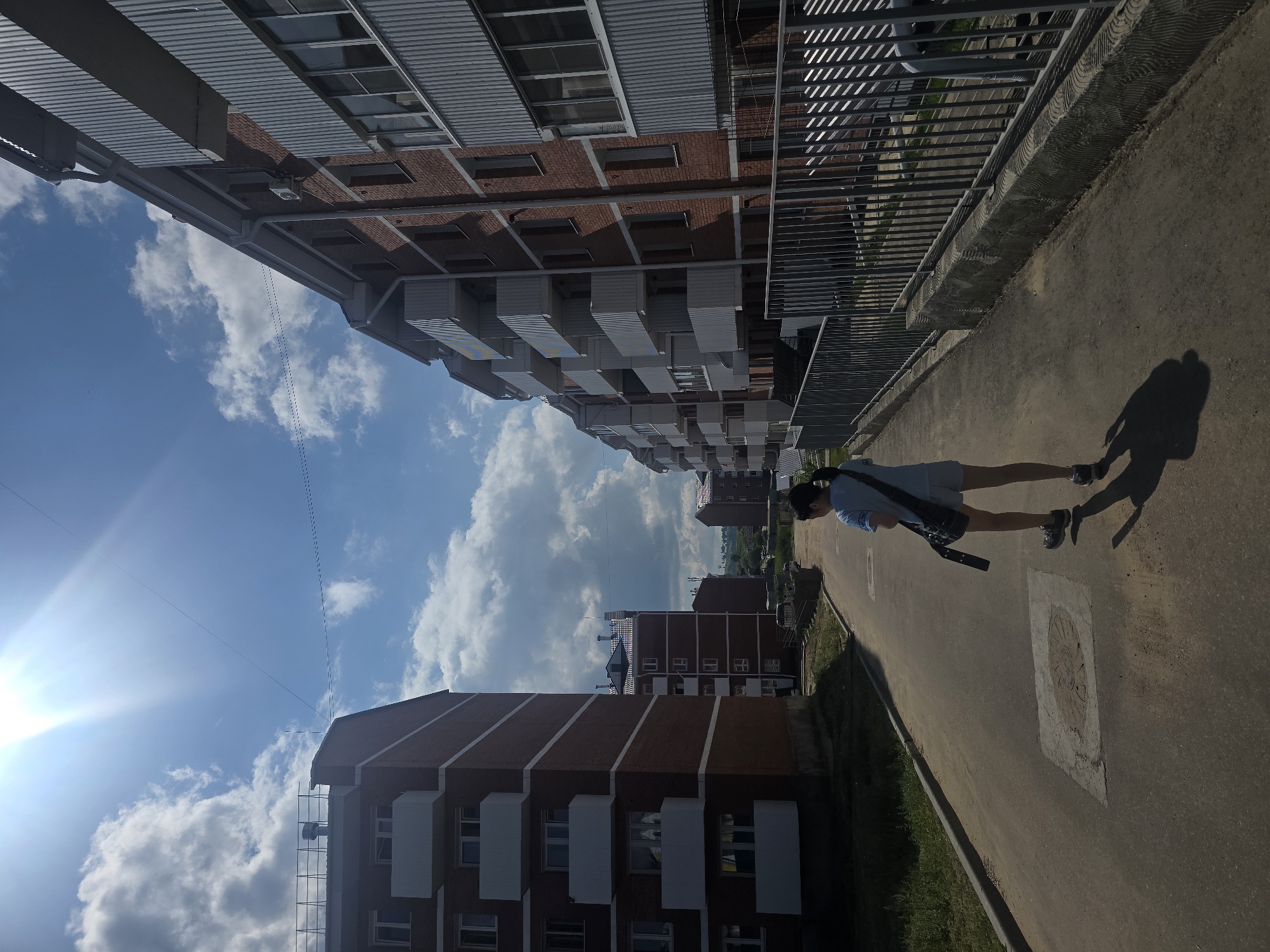 A young female with dark hair, wearing a light blue t-shirt and grey shorts, walks away from the camera down a paved road. She has a dark crossbody bag slung over her right shoulder. She is wearing athletic shoes. The sun is bright and high in the sky, casting a long shadow of the woman on the road. On the left side of the road, there is a multi-story brick apartment building with white balconies. On the right side of the road, there is a row of similar apartment buildings with red brick and white balconies, bordered by a metal fence. The sky is blue with scattered white clouds. The scene appears to be in a residential area of Ulan-Ude, Russia, likely a new development given the modern apartment buildings.
