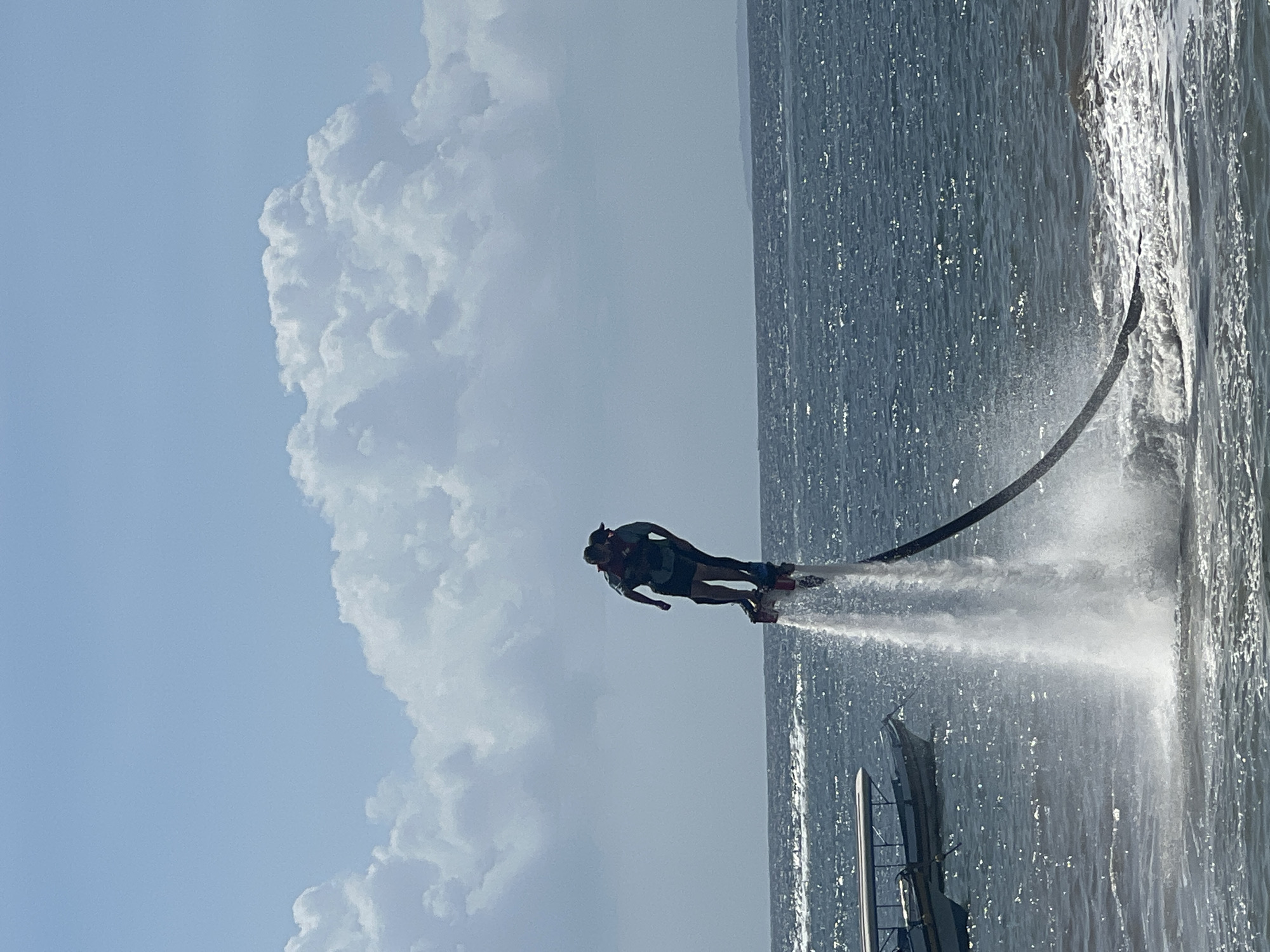 In Kuta Selatan, Indonesia, two individuals are engaged in a watersport activity, propelled into the air by a hydro-powered device. One individual is positioned behind the other, with both wearing life vests, suggesting a tandem ride on a flyboard or similar equipment. They are suspended approximately fifteen feet above the dark blue, choppy ocean, with a powerful jet of water erupting from beneath their feet, creating a misty spray. A black hose, the conduit for the water, extends from the device downwards and out of frame, presumably connected to a watercraft. In the mid-ground, to the left, two traditional Indonesian fishing boats with white canopies are anchored on the water. The horizon line is visible, separating the sea from a hazy, distant landmass. Above, the sky is a clear, pale blue, punctuated by large, fluffy white cumulus clouds. The overall scene captures a moment of exhilaration and outdoor recreation in a tropical marine environment.