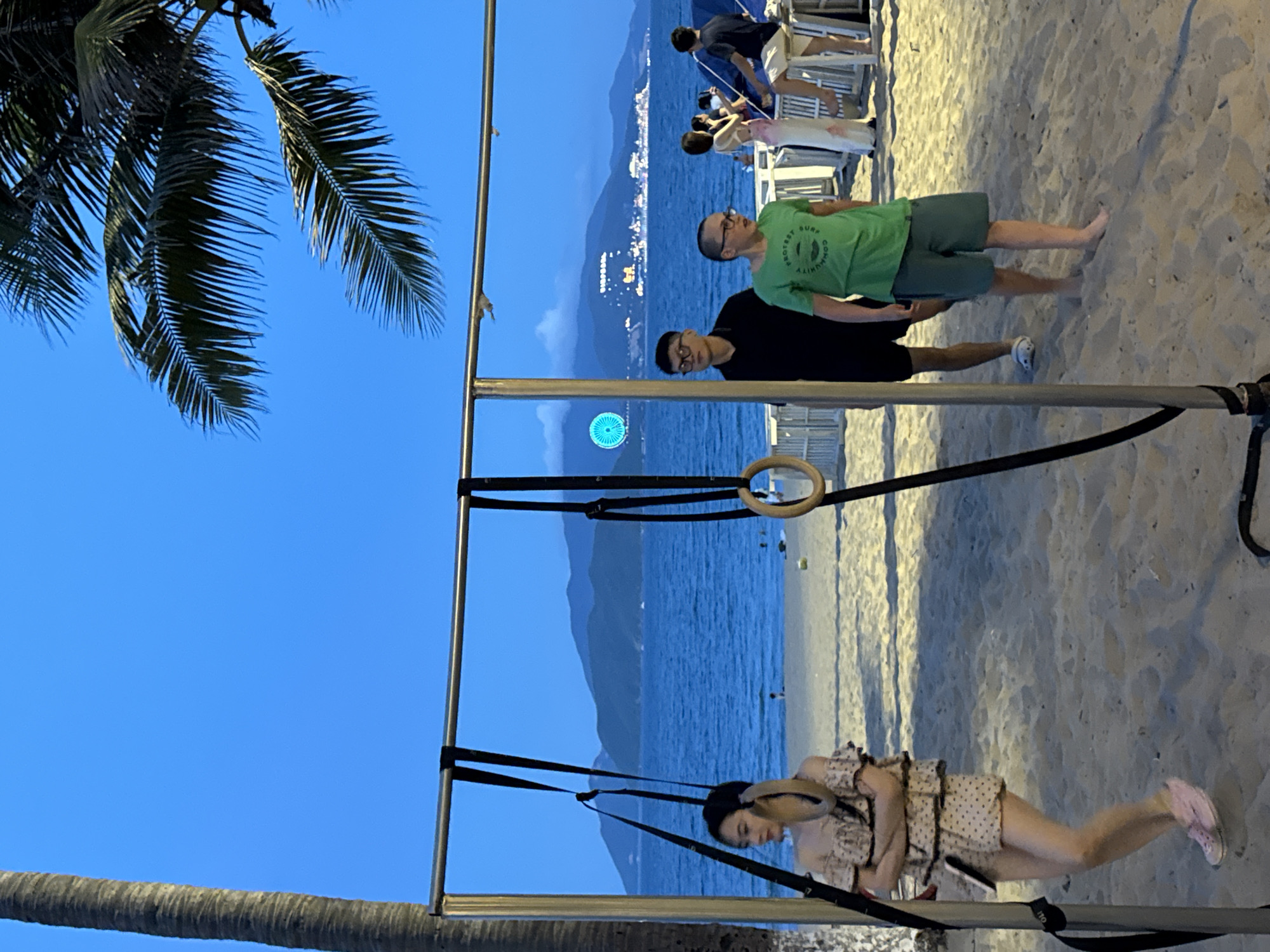 An evening scene unfolds on a sandy beach in Nha Trang, Vietnam, with a calisthenics rig as a central element. The sky is a deep twilight blue, transitioning from the last remnants of daylight. A large palm tree trunk stands on the left, and its fronds are visible against the sky at the top right.
In the foreground, a young woman stands on the sand to the left, wearing a ruffled off-the-shoulder top and a patterned skirt. She has her arms crossed and is looking to her left. To her right, two young men are walking on the sand. The first, wearing glasses and a black short-sleeved shirt and shorts, is partially obscured. The second, also wearing glasses, is in a green t-shirt and green shorts.
The calisthenics rig, a metal frame with parallel bars, extends across the middle of the image. Gymnastic rings hang from black straps attached to the top bar.
In the mid-ground, the calm blue ocean stretches to the horizon, meeting a range of hazy mountains. Scattered across the beach are white beach chairs and a few other figures, some of whom appear to be seated or standing near a blue tent-like structure. In the distance, lights from a coastal city are visible on the mountainside, and a Ferris wheel is illuminated on the left side of the frame. The sand itself is a warm, pale color, showing subtle undulations and footprints.