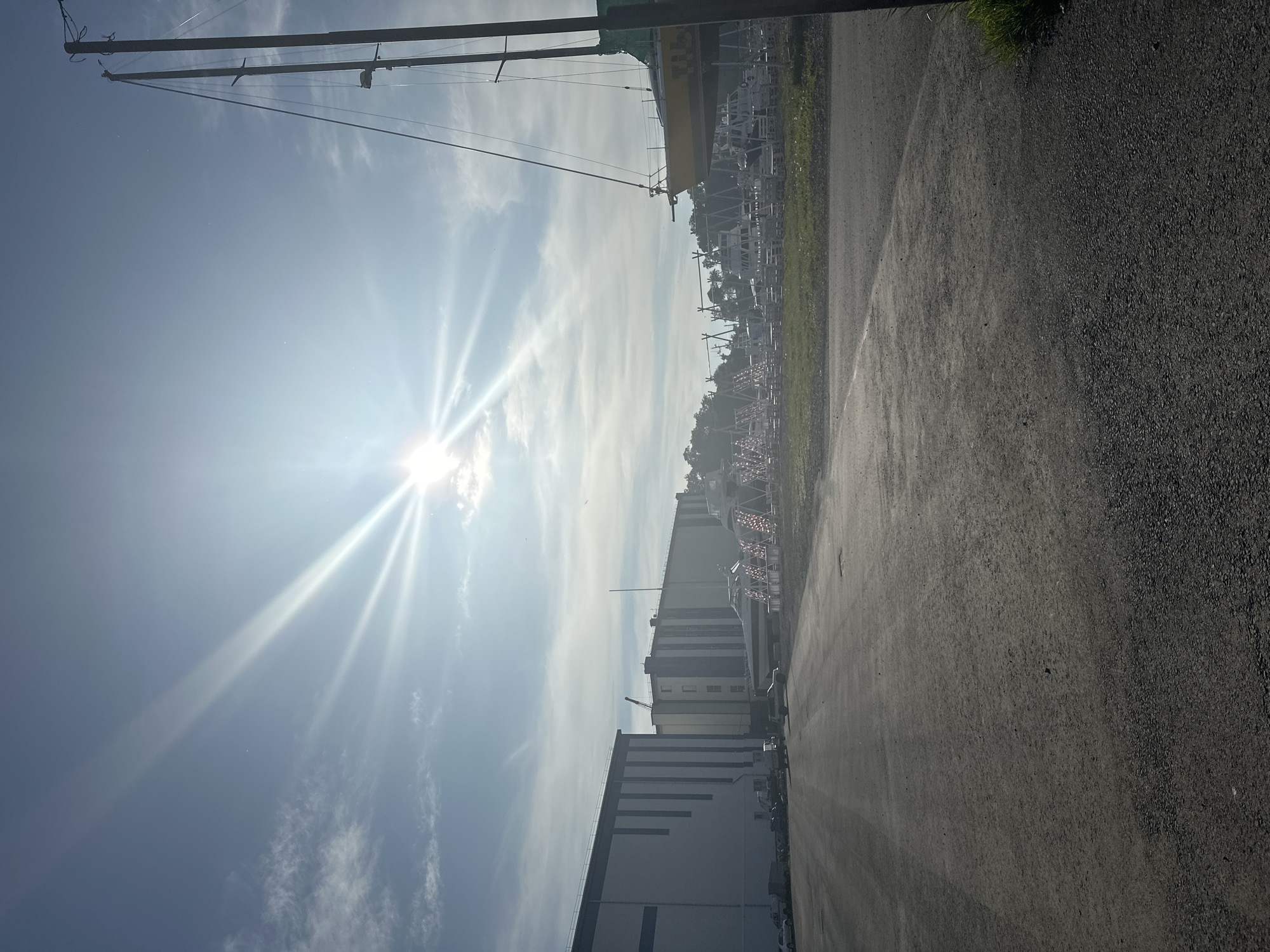 The image depicts an outdoor scene with a bright sun shining through a partly cloudy sky, casting lens flares. In the foreground, a paved area transitions to a grassy area. In the midground, several boats are visible, some on dry land and on stands, and a large, modern building complex is situated to the left. To the right, the mast and rigging of a sailboat are prominent against the sky. The overall impression is of a shipyard or marina. The location is specified as Petropavlovsk-Kamchatsky, Russia.